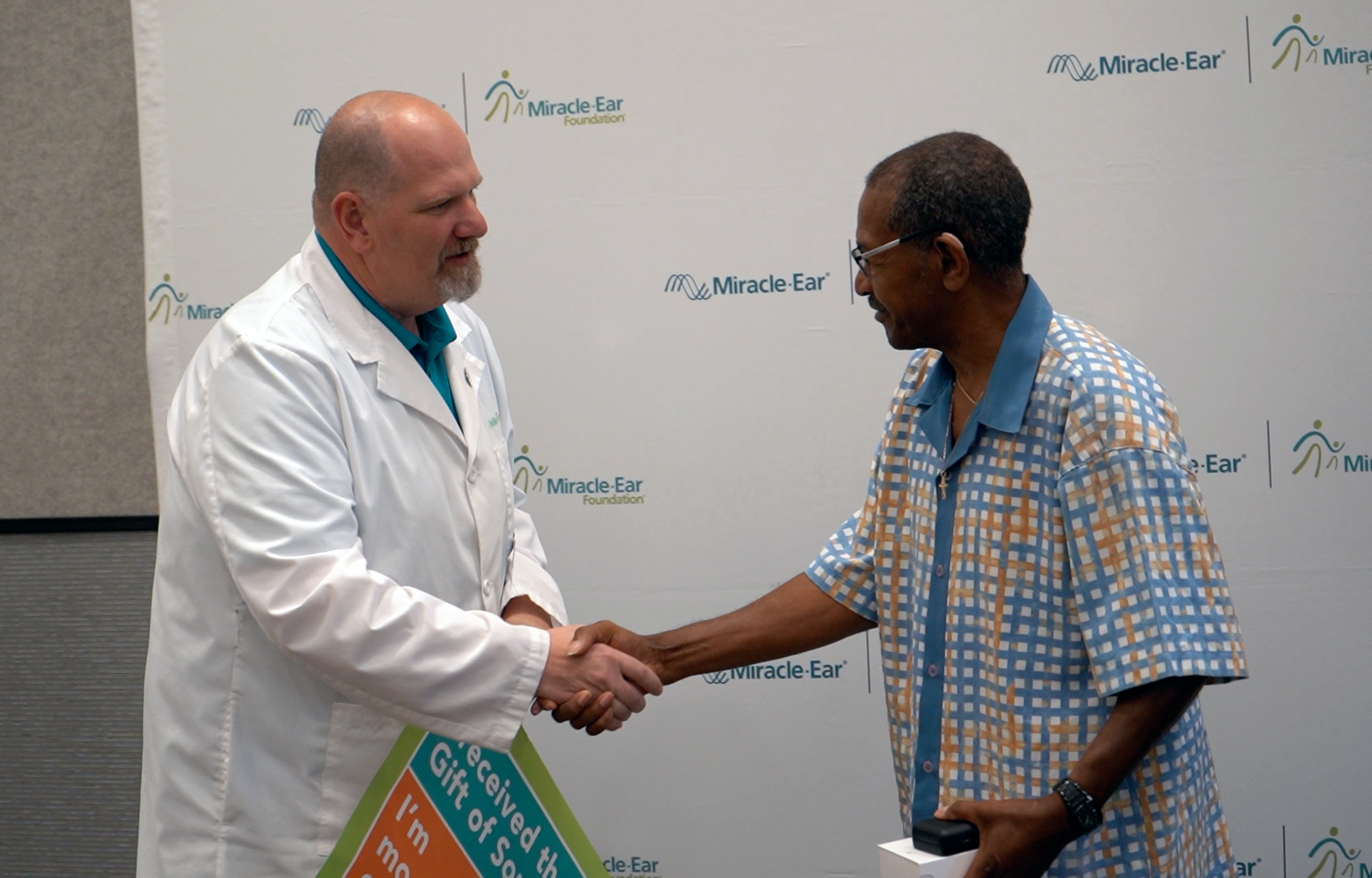 A hearing specialist shakes hands with a recipient in front of a Miracle-Ear backdrop