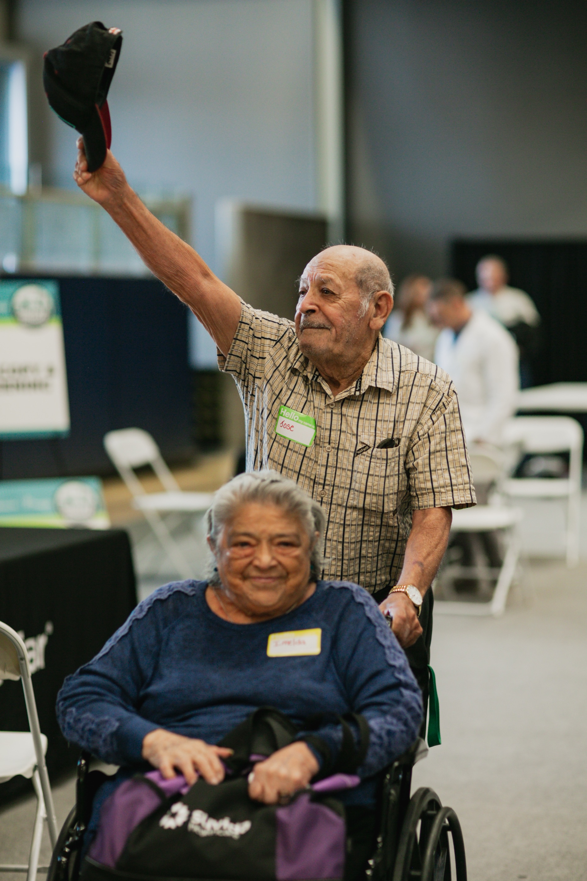 A man raises his hat in celebration behind a smiling woman seated in a wheelchair