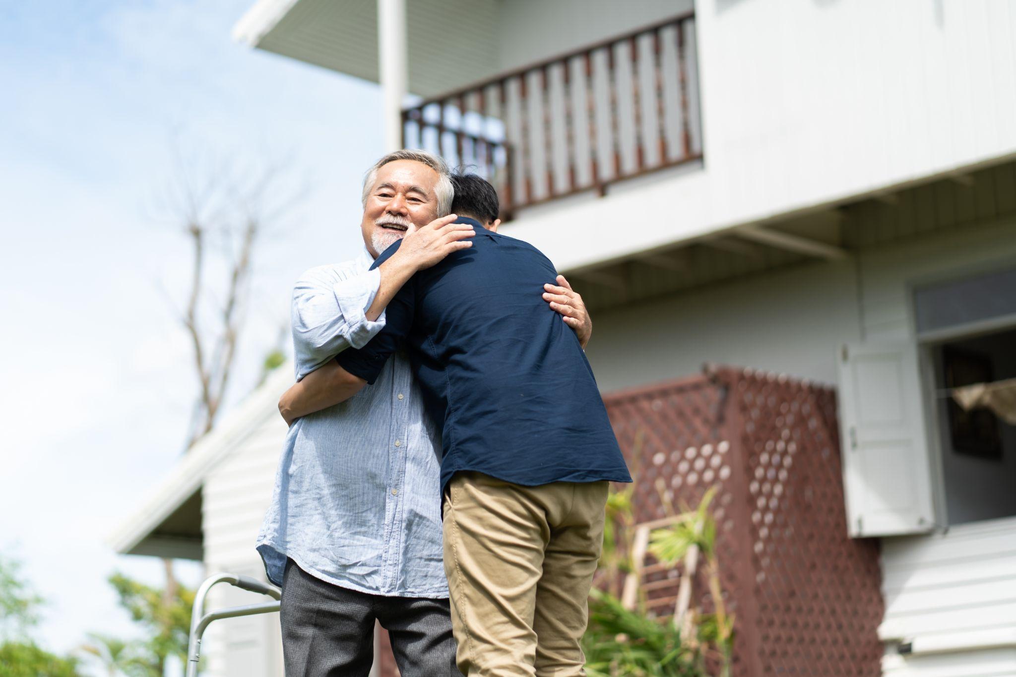 An older man embraces a younger man outside a house, a walking frame beside them.