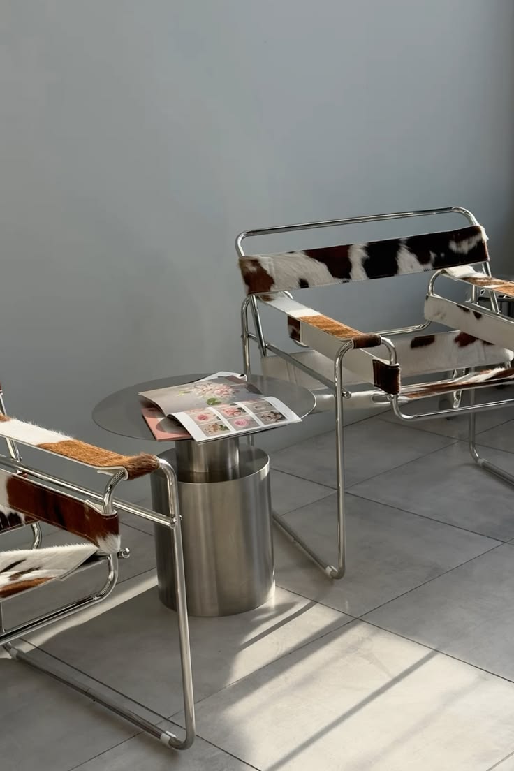 Two modern metal chairs with cowhide upholstery next to a metal round table holding an open magazine.