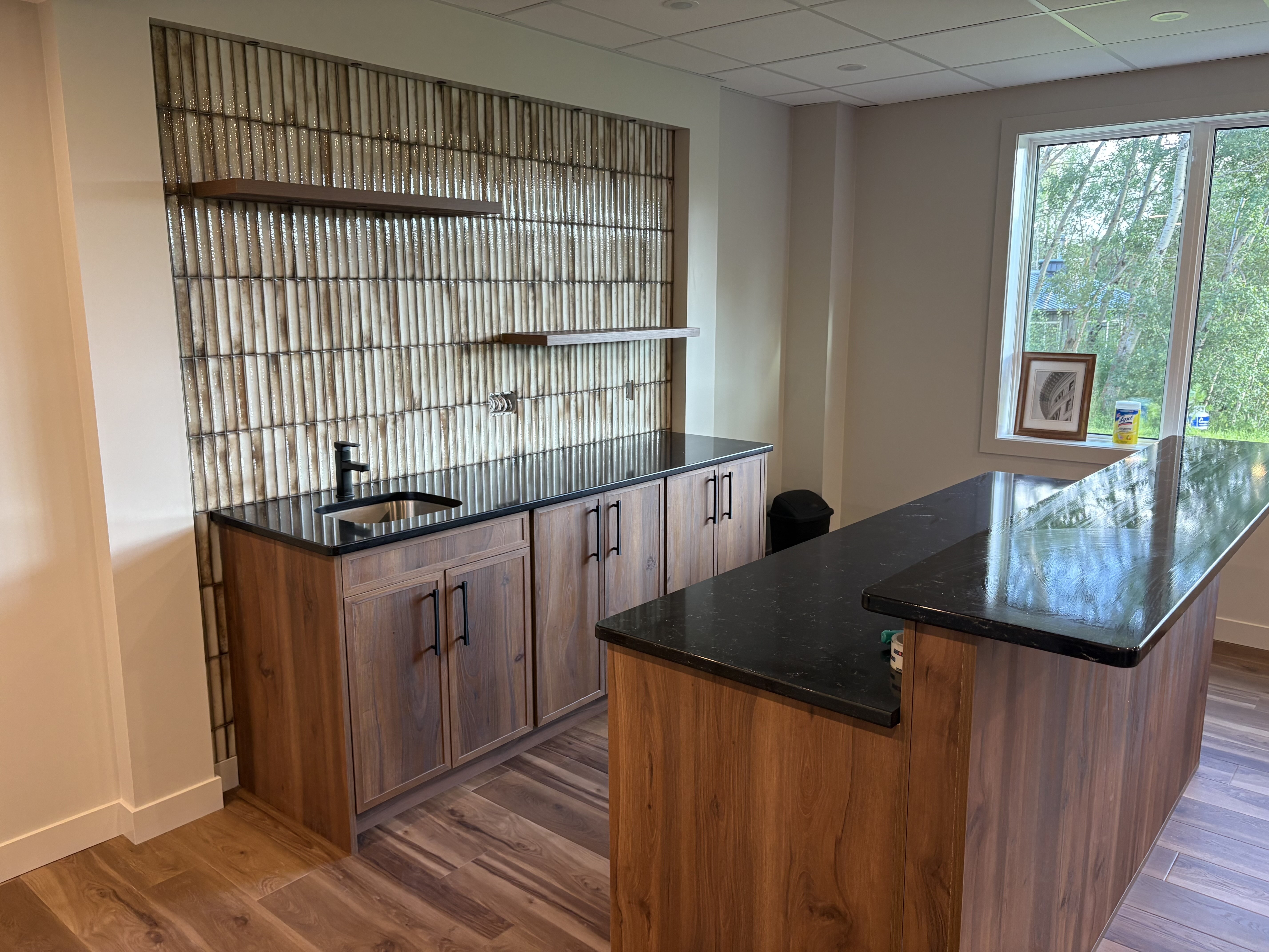 Custom basement wet bar with warm wood cabinetry, dark countertops, seating peninsula and full-height textured tile feature wall.