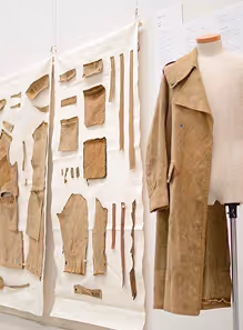 Tailored beige trench coat displayed on a mannequin next to fabric pattern pieces pinned to a board.