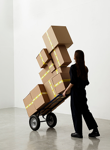 Person in black clothes moving a hand truck stacked high with cardboard boxes sealed with yellow tape.
