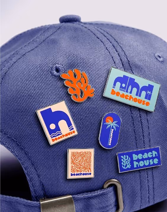Blue baseball cap decorated with six colorful enamel pins featuring beach-themed designs and the word 'beachouse'.