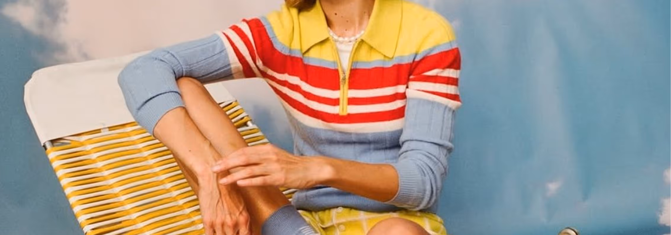 Person in colorful striped sweater and yellow checkered shorts sitting with legs crossed on a yellow-striped chair against a blue and beige background.