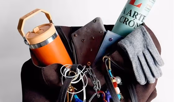 Top view of a brown bag filled with a rolled newspaper, orange thermos, grey gloves, and various keychains and accessories.