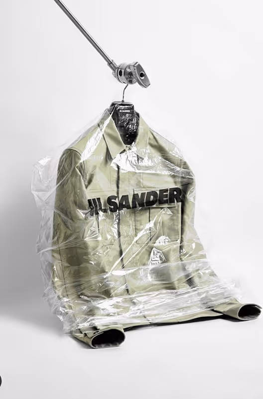 Light green Jil Sander jacket covered in a clear plastic garment bag hanging from an industrial clamp.