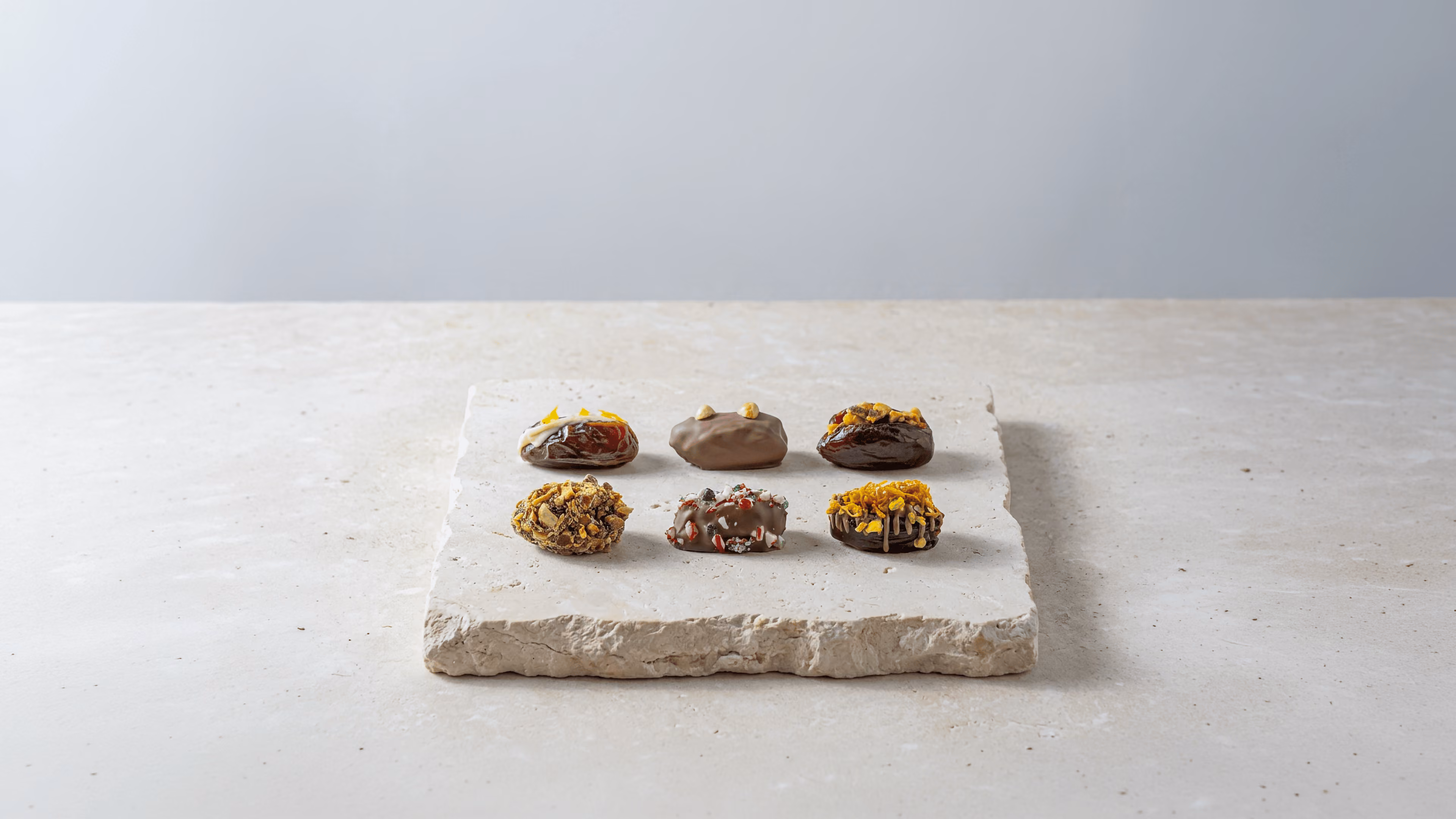 Six stuffed Medjool dates arranged on a stone plate beside a glass of tea on a neutral stone surface.
