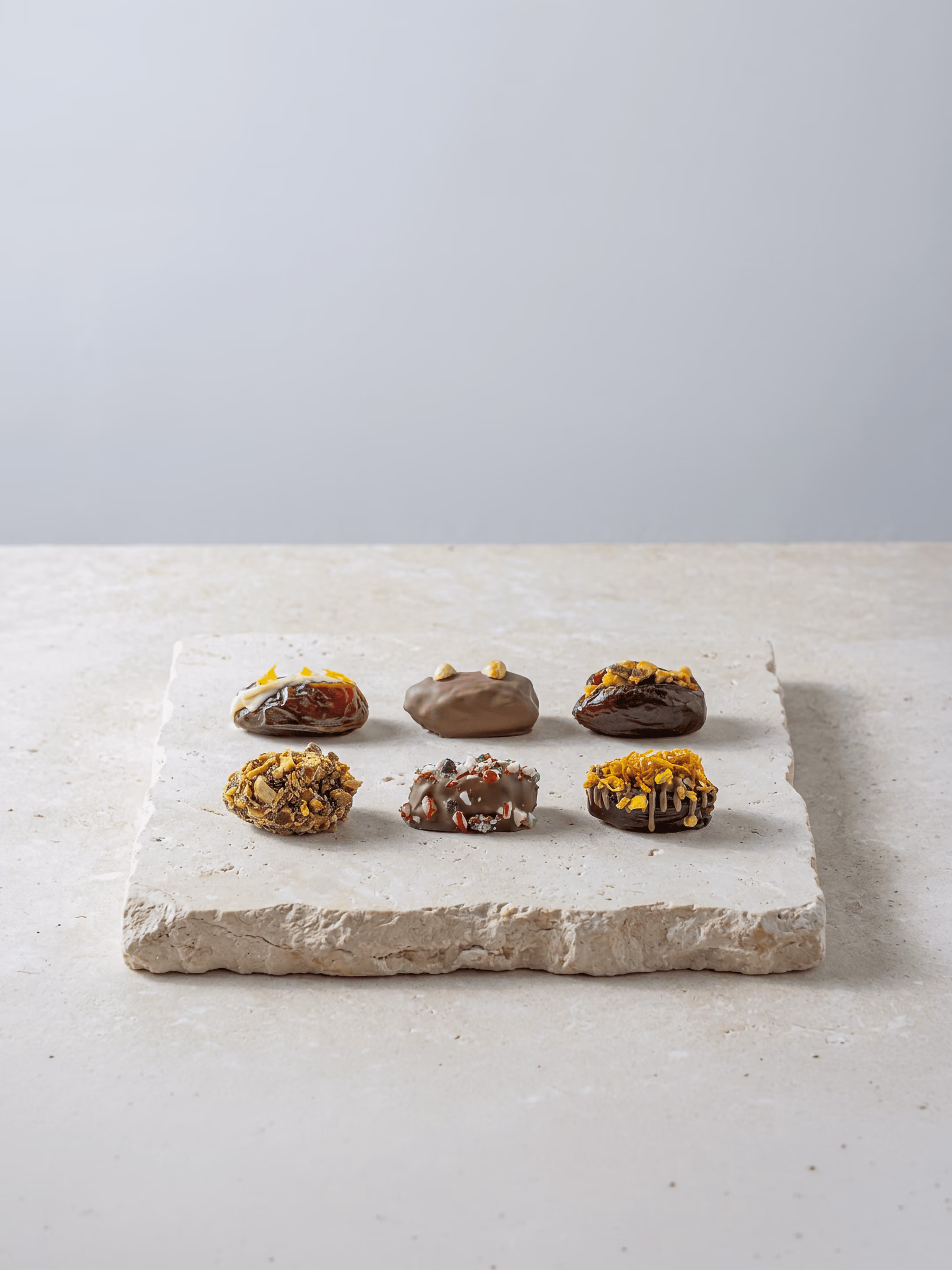 Six stuffed Medjool dates arranged on a stone plate beside a glass of tea on a neutral stone surface.
