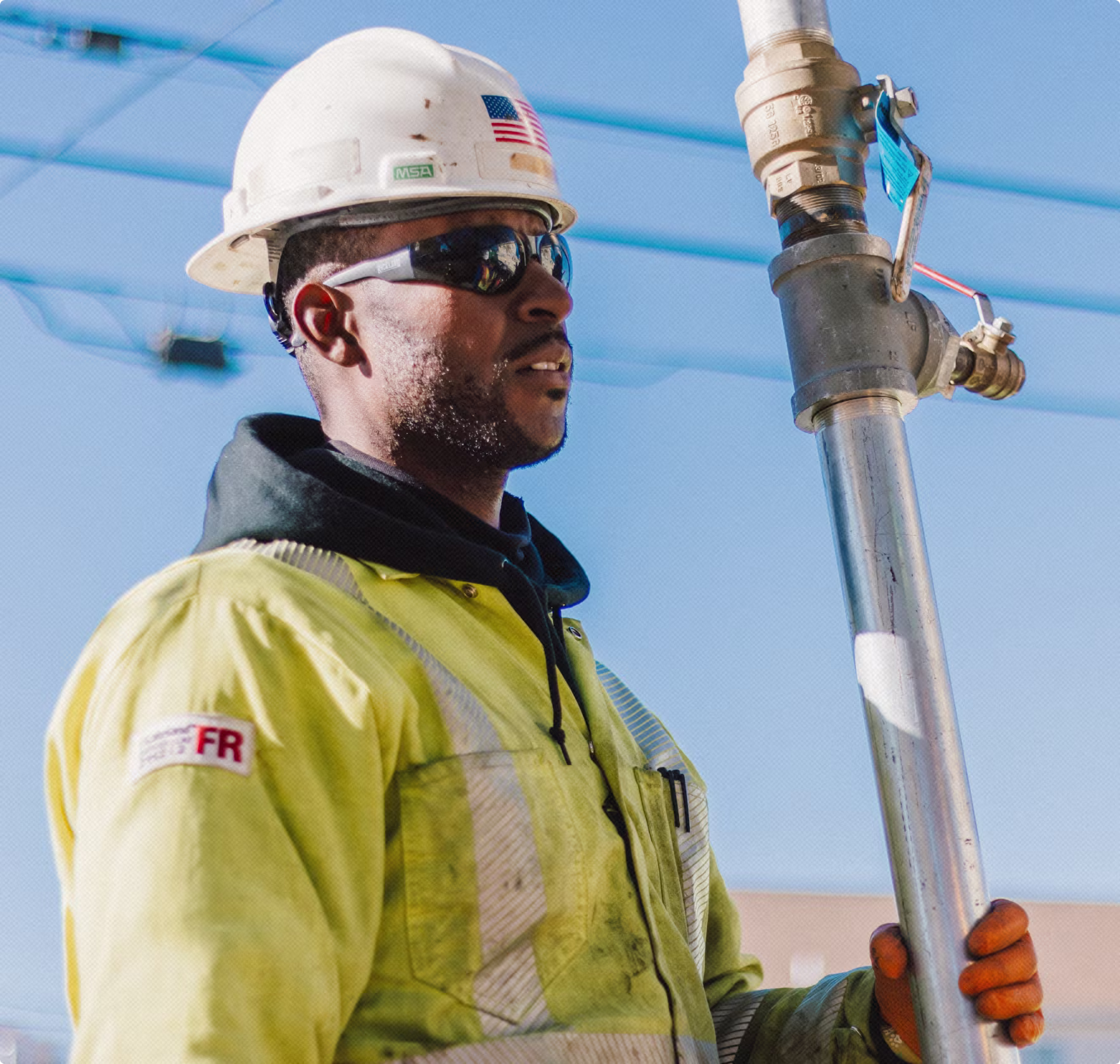 Construction worker wearing a hard hat and safety glasses holding a large metal pipe outdoors.