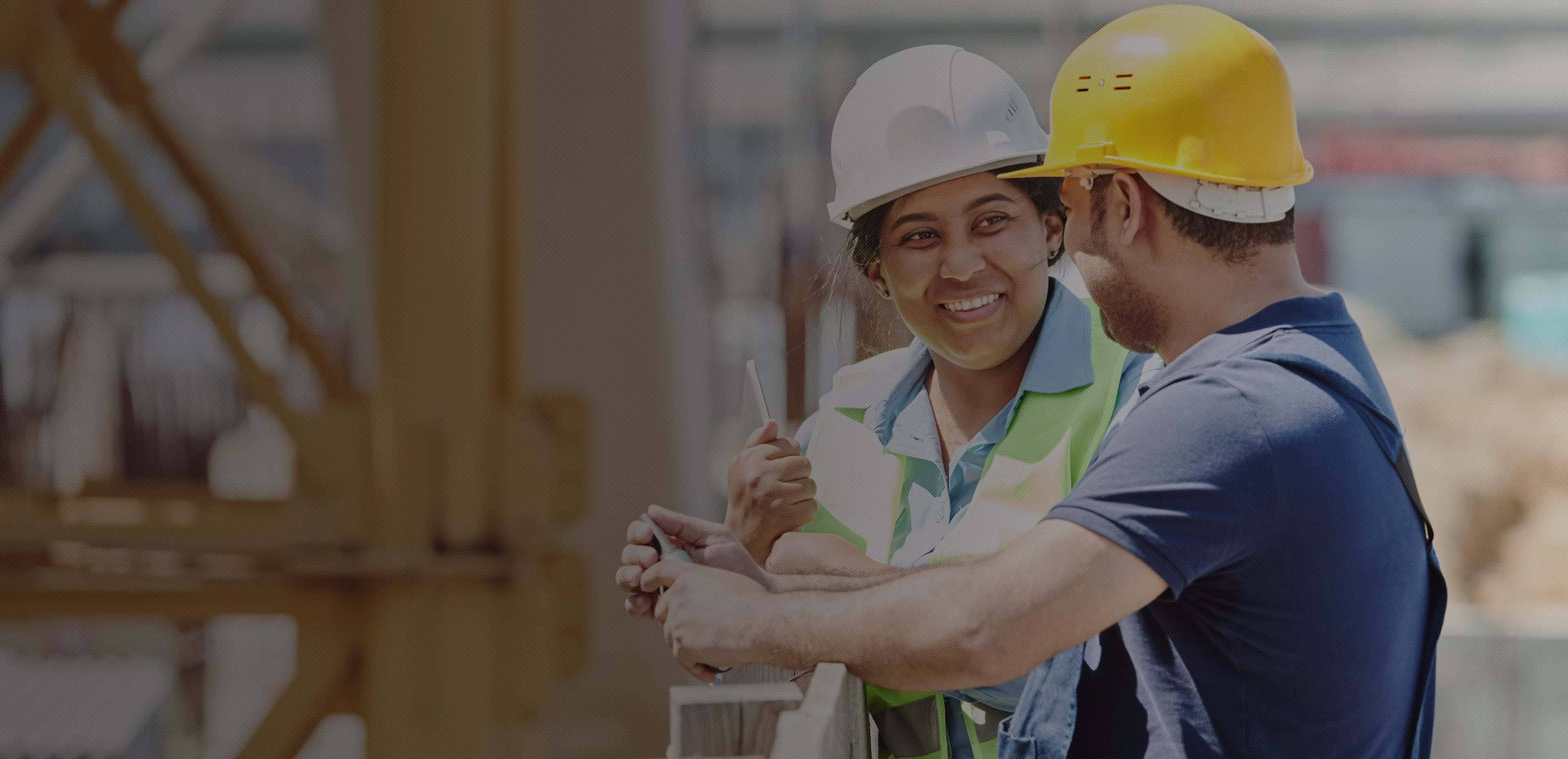 Two construction workers wearing safety helmets and vests smiling and talking at a construction site.