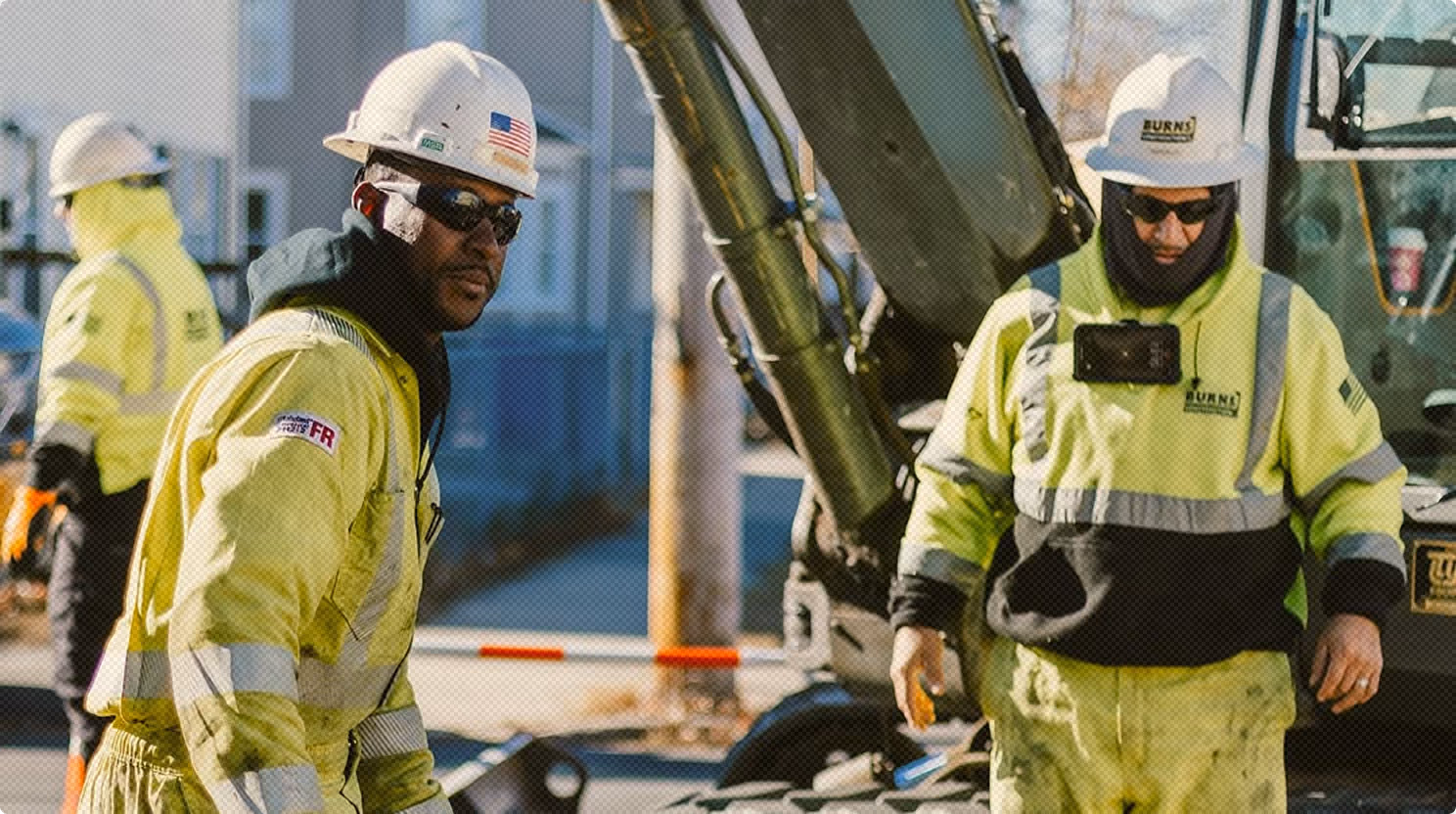 Two construction workers wearing high-visibility yellow jackets, hard hats, and sunglasses standing at a worksite with machinery in the background.