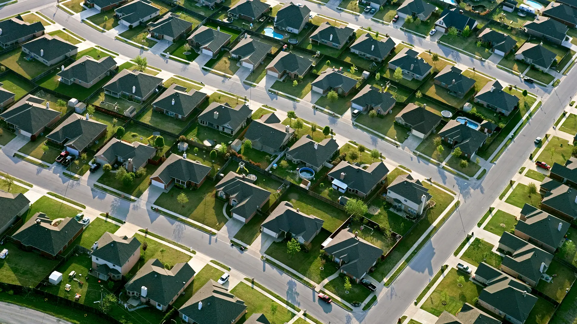 Aerial view of a residential neighborhood with rows of detached homes, wide intersecting streets, green lawns, fenced backyards, driveways, and scattered backyard pools. The homes are arranged in a grid pattern and photographed in warm daylight from above.