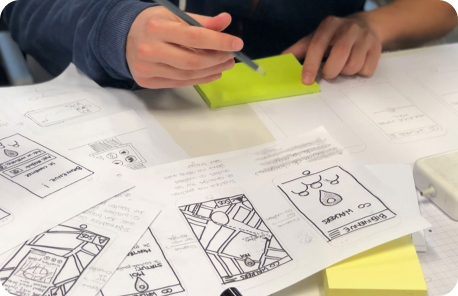 Product designer working with low-fidelity wireframes of a mobile app on a table. They are holding a pen and a stack of sticky notes.