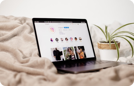 A laptop placed on a bed next to a potted plant. On the laptop screen is the web version of instagram.