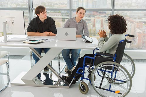 Three people working together at a desk in an office, one person in a wheel chair
