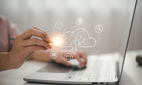 A close up of a person working at a laptop with one hand holding a stylus and tapping a graphic overlay of a cloud and other icons branching out from it.