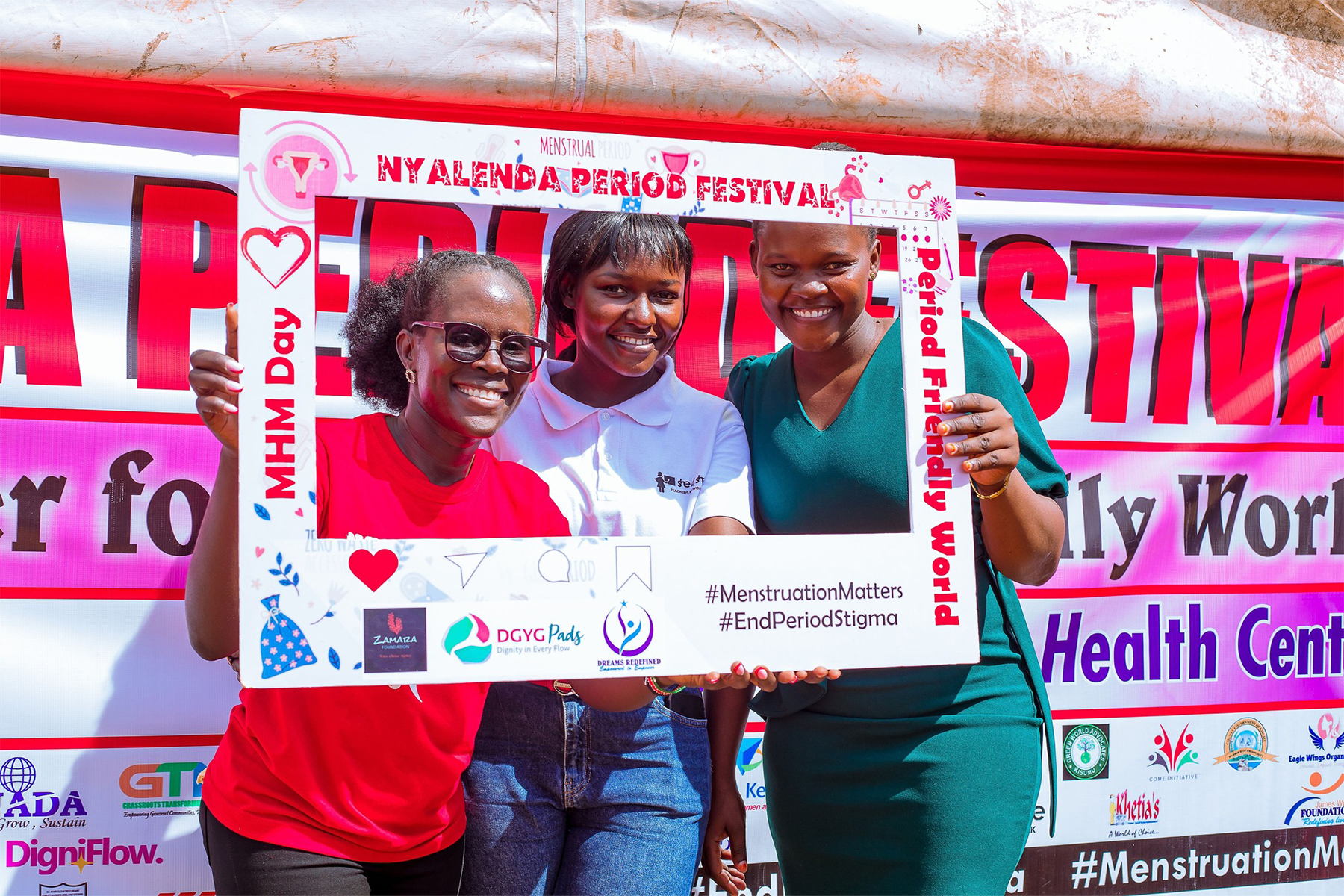 Three women pose behind a large picture frame in front of a poster for the Nyalenda Period Festival.