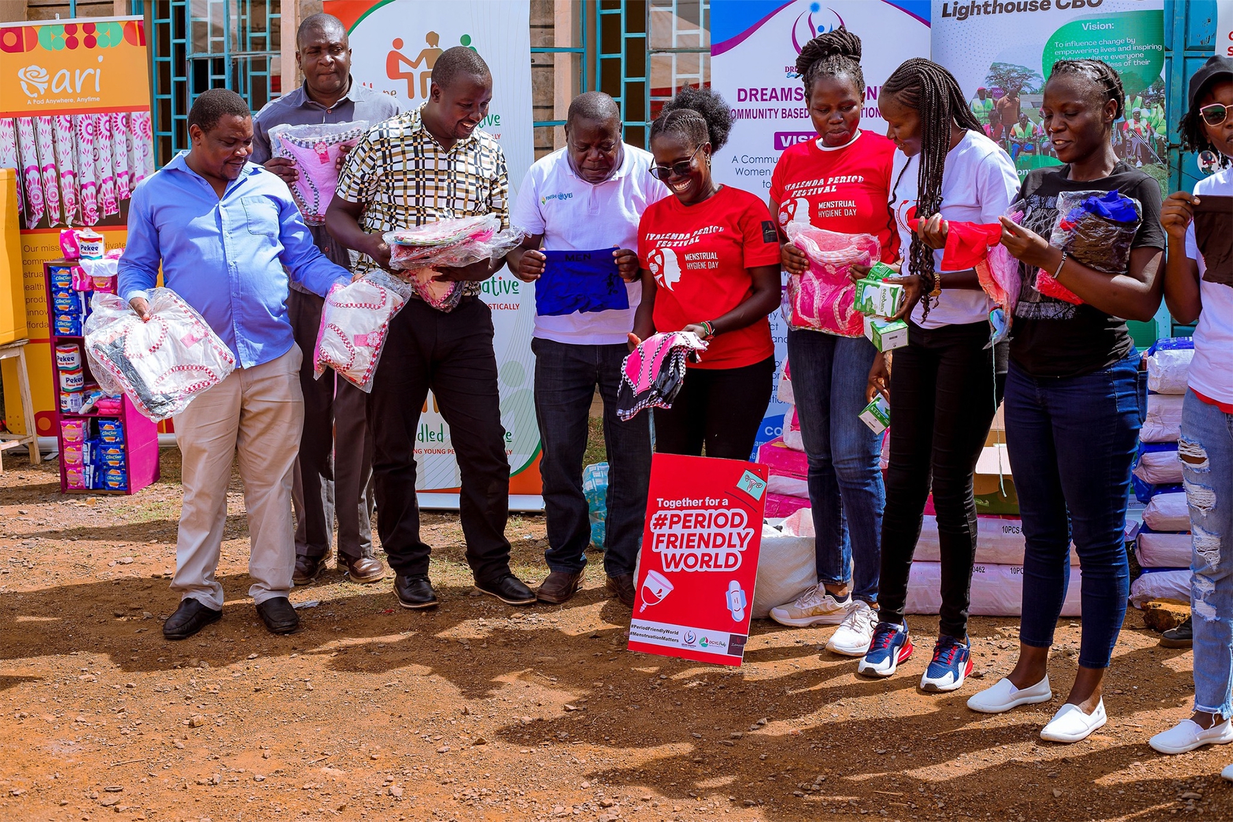 A group of people holds period products and laughs. To their feet is a poster that says "Together for a #PeriodFriendlyWorld.