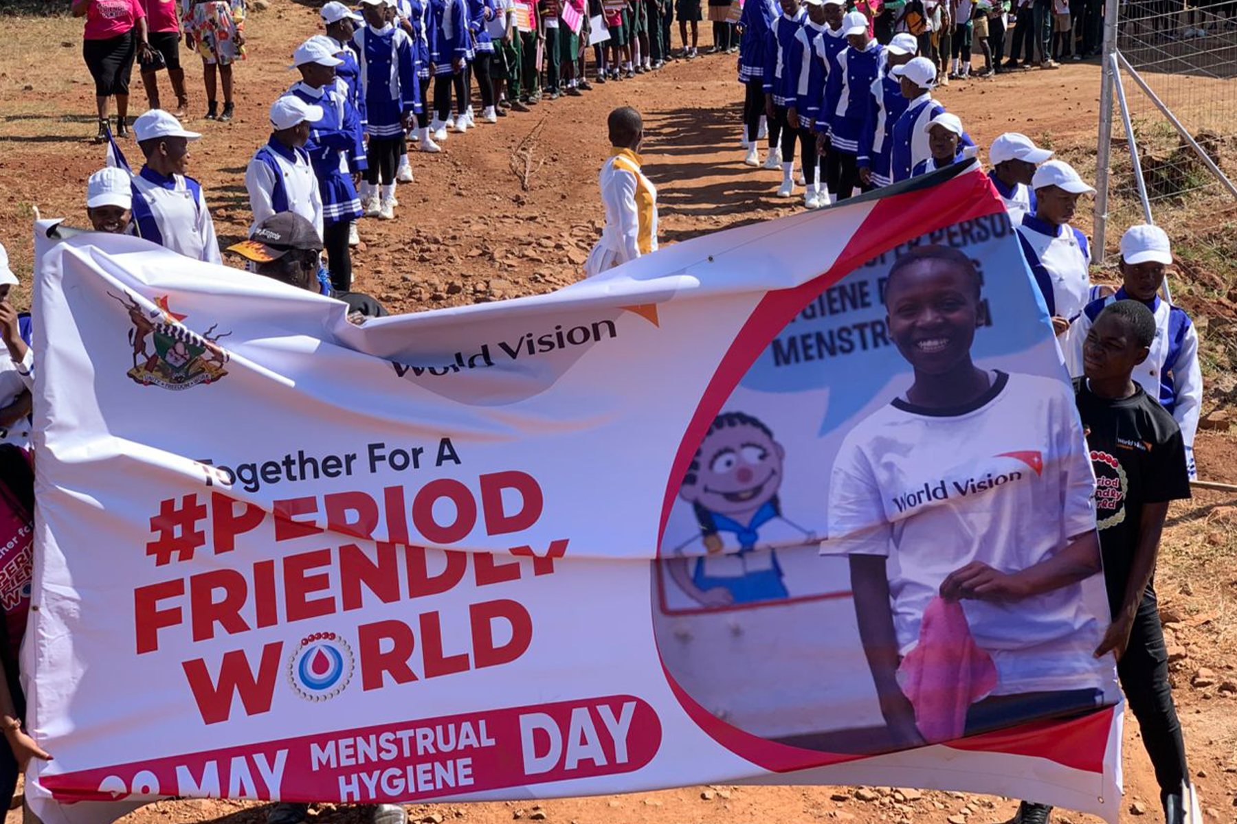 A big banner saying "Together for a #PeriodFriendlyWorld is held up in front of many people that stand in two lines to form a path.