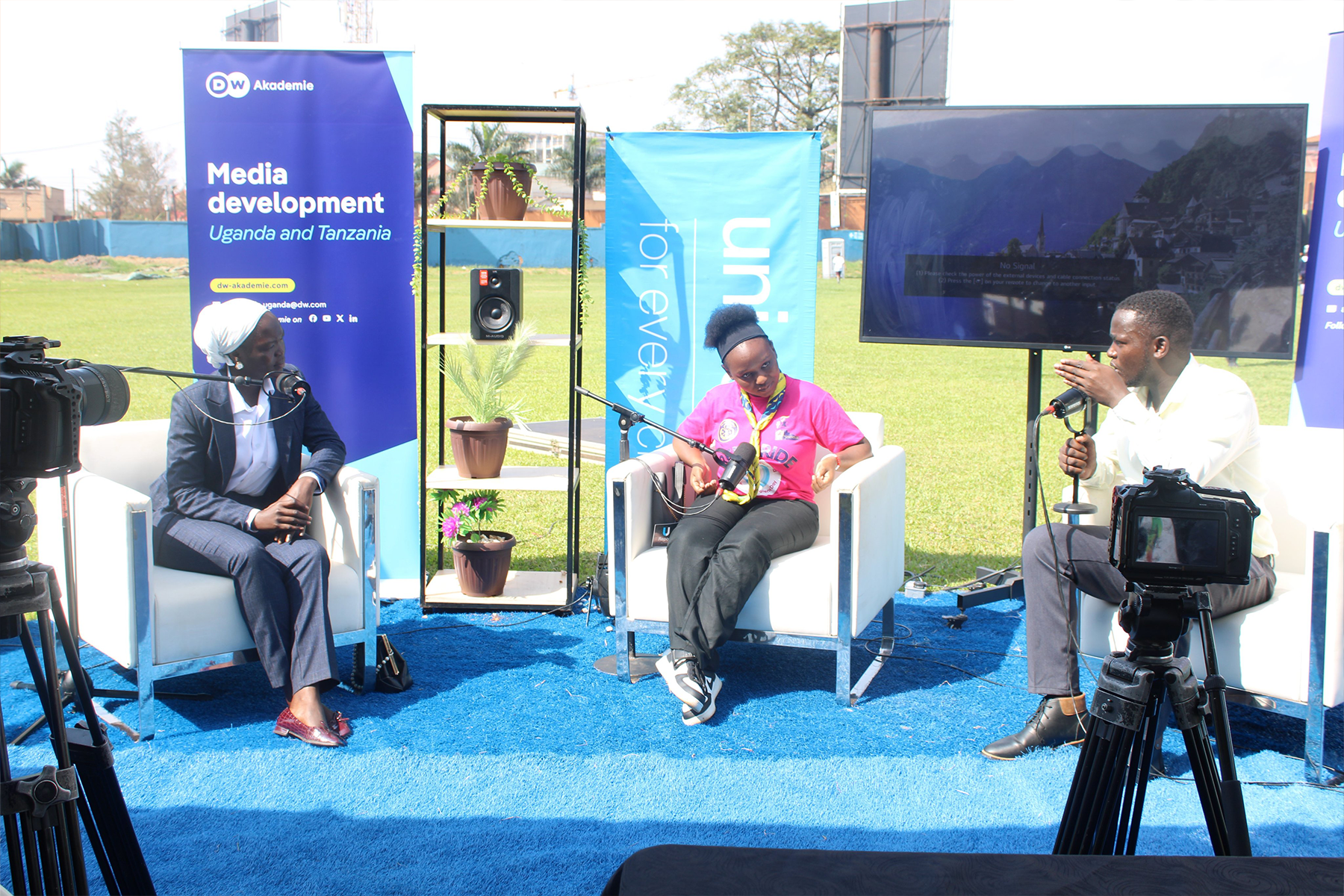 Three people sit on a blue stage with banners and a screen and speak into microphones.