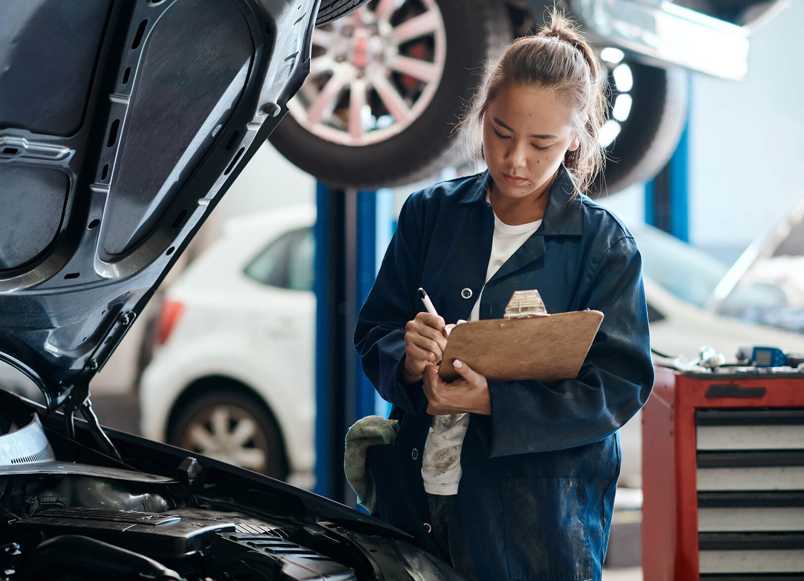 Mechanical woman writing notes on a pad, beside a car hood.