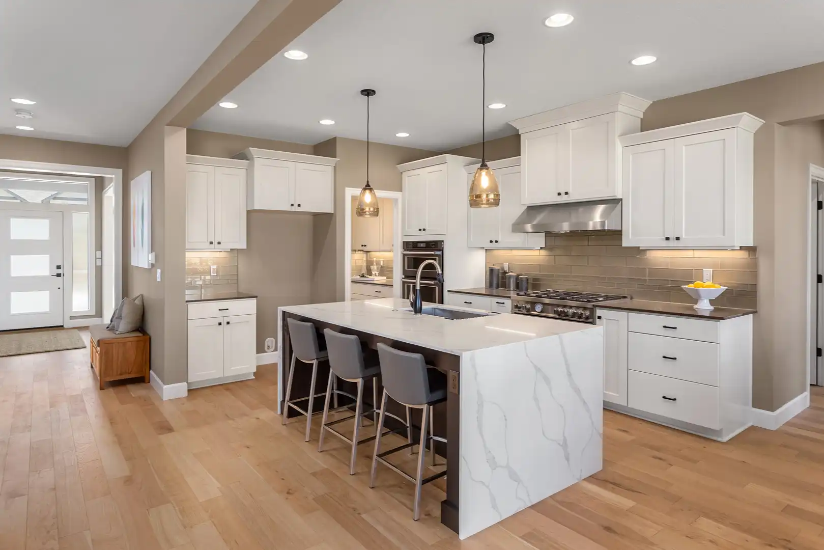 Modern kitchen with white cabinets, a marble island with sink, three gray bar stools, and pendant lights.