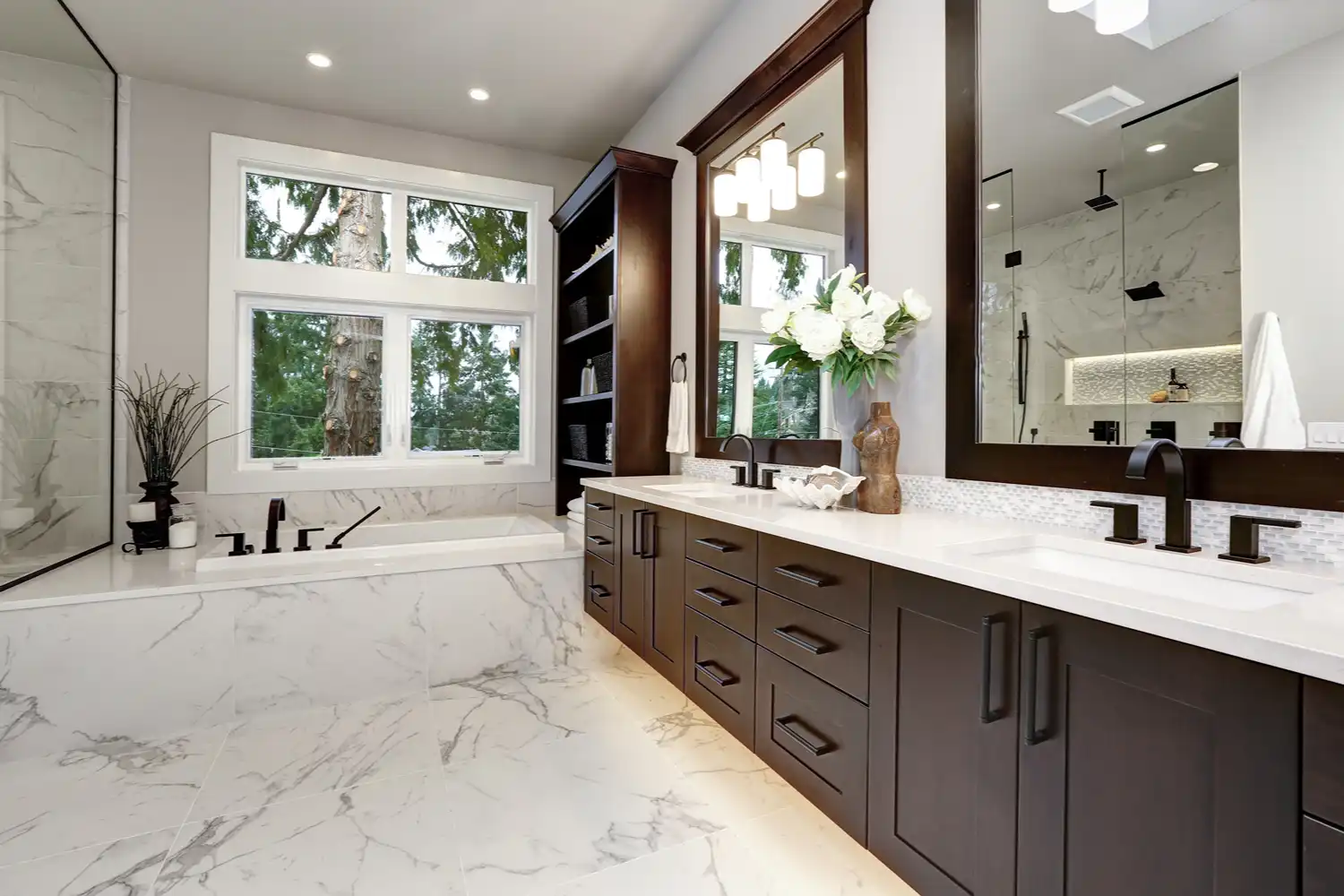 Modern bathroom with dark wood vanity, large mirrors, white marble floors, and built-in bathtub by window.