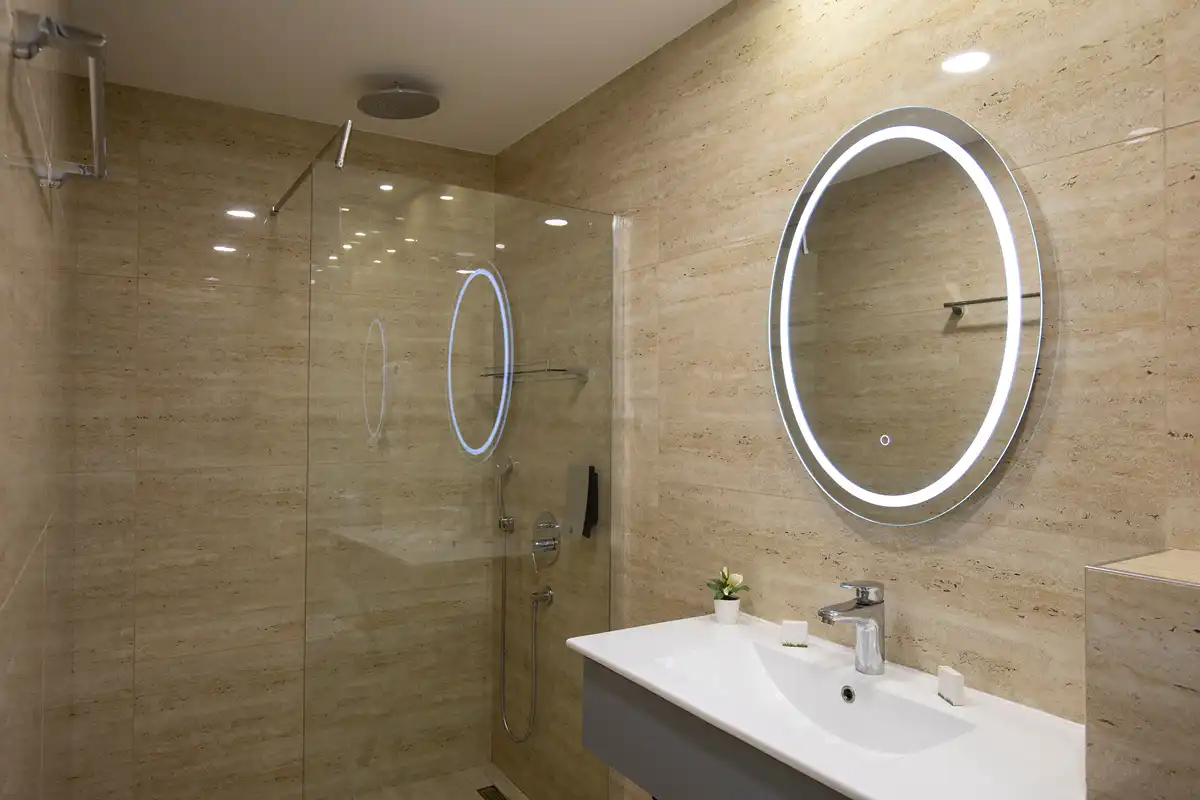 Modern bathroom with round illuminated mirror, white sink, and glass shower enclosure with beige tile walls.