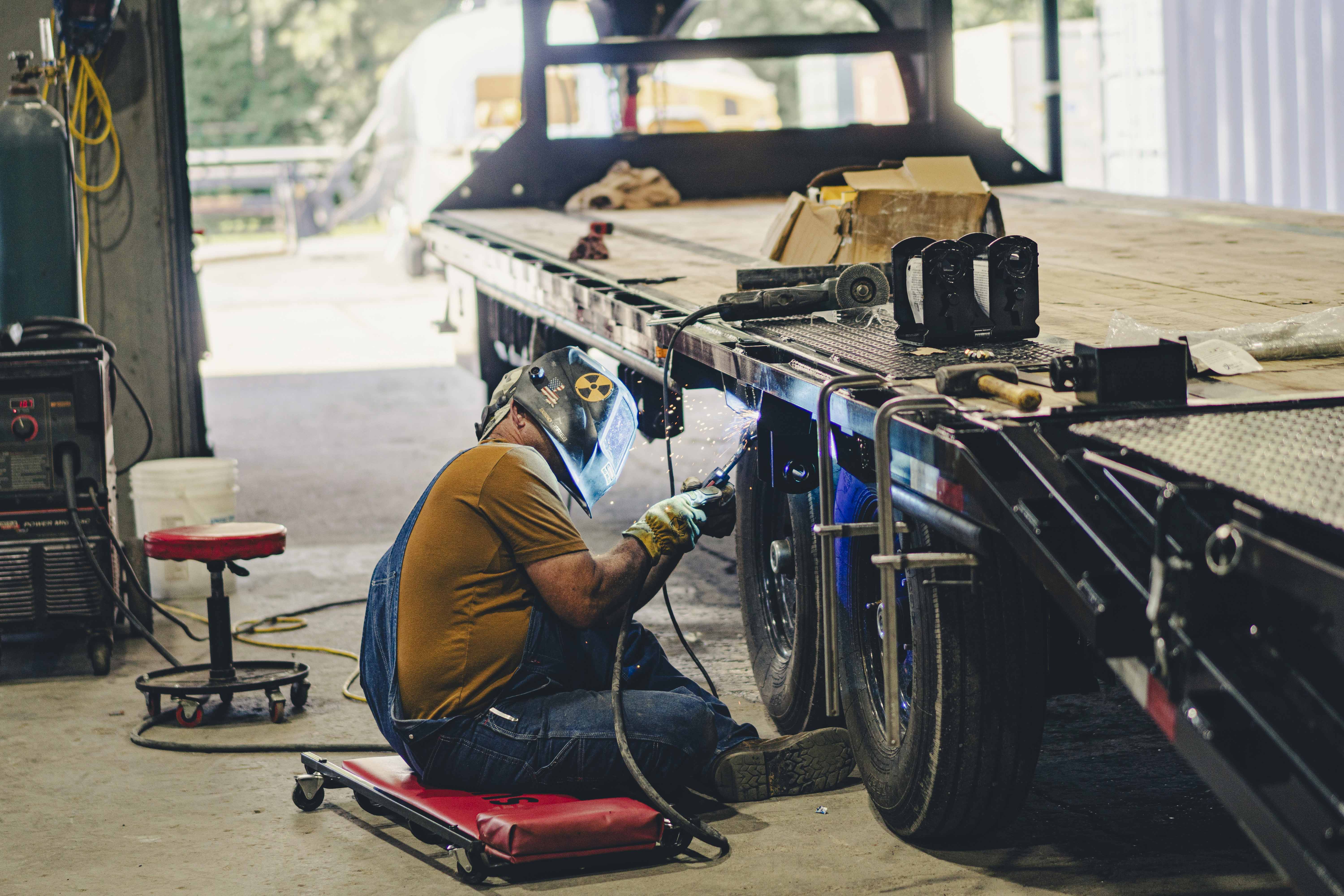 A man wearing a welding helmet, gloves, and overalls is seated on a red creeper, welding the underside of a flatbed trailer in a workshop. Sparks fly from the welding point, and tools and materials are scattered on top of the trailer. The background includes welding equipment, a stool, and open garage doors with light streaming in.