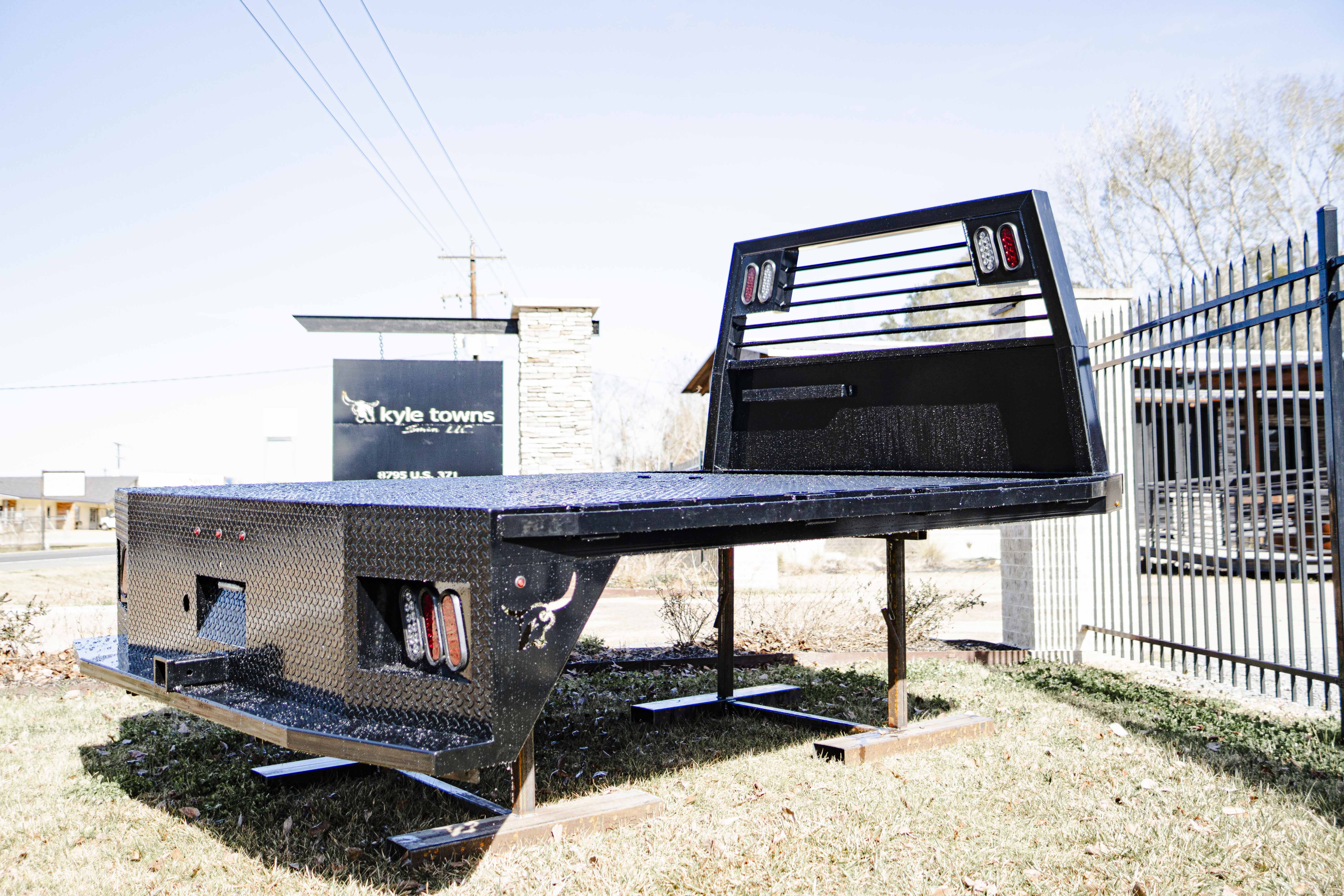 Black metal flatbed truck bed with diamond plate surface displayed outdoors on grass near a gated fence.