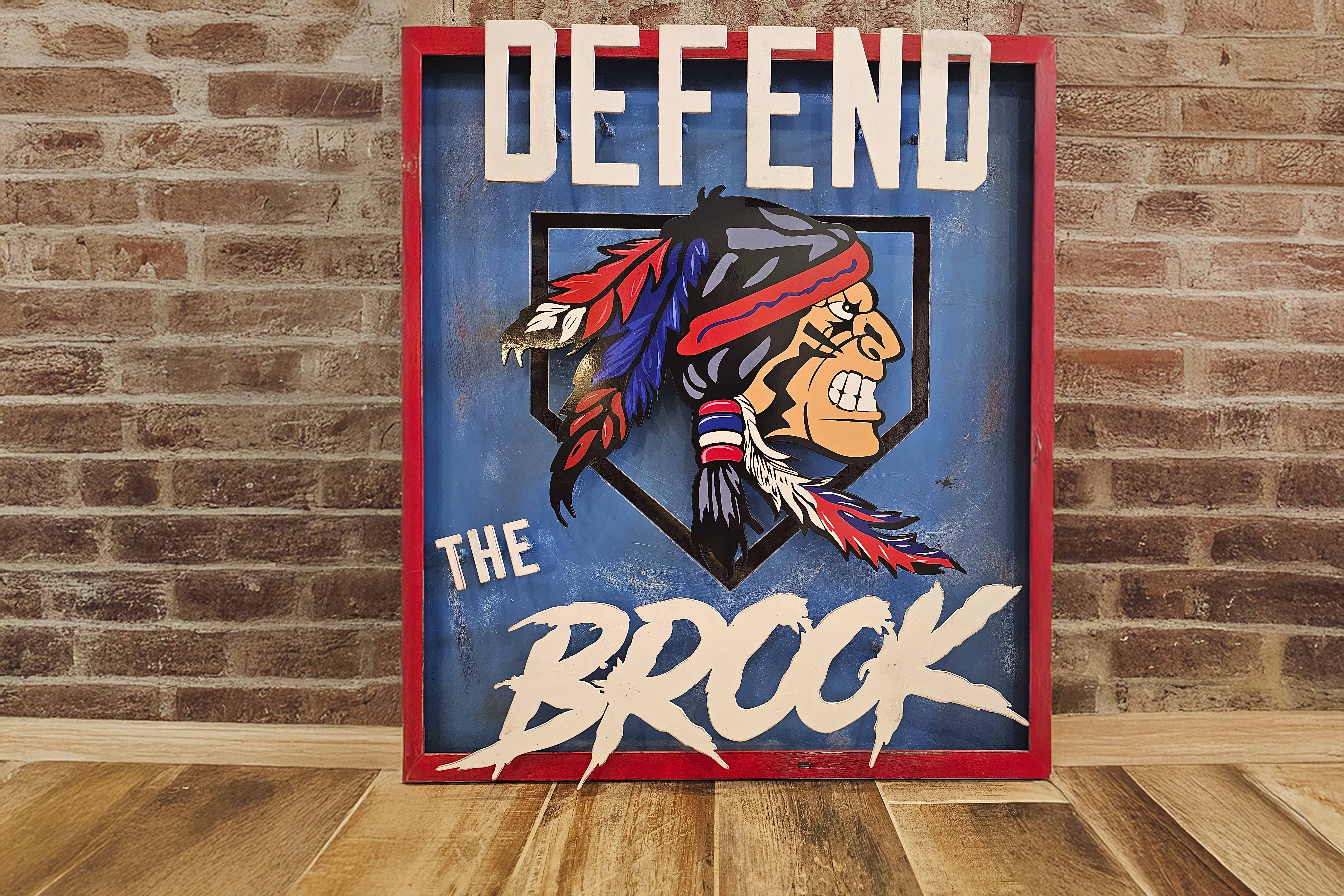 Framed sign with an angry Native American mascot head with feathers and the text ‘DEFEND THE BROOK’ on a blue background, placed on a wooden floor in front of a brick wall.