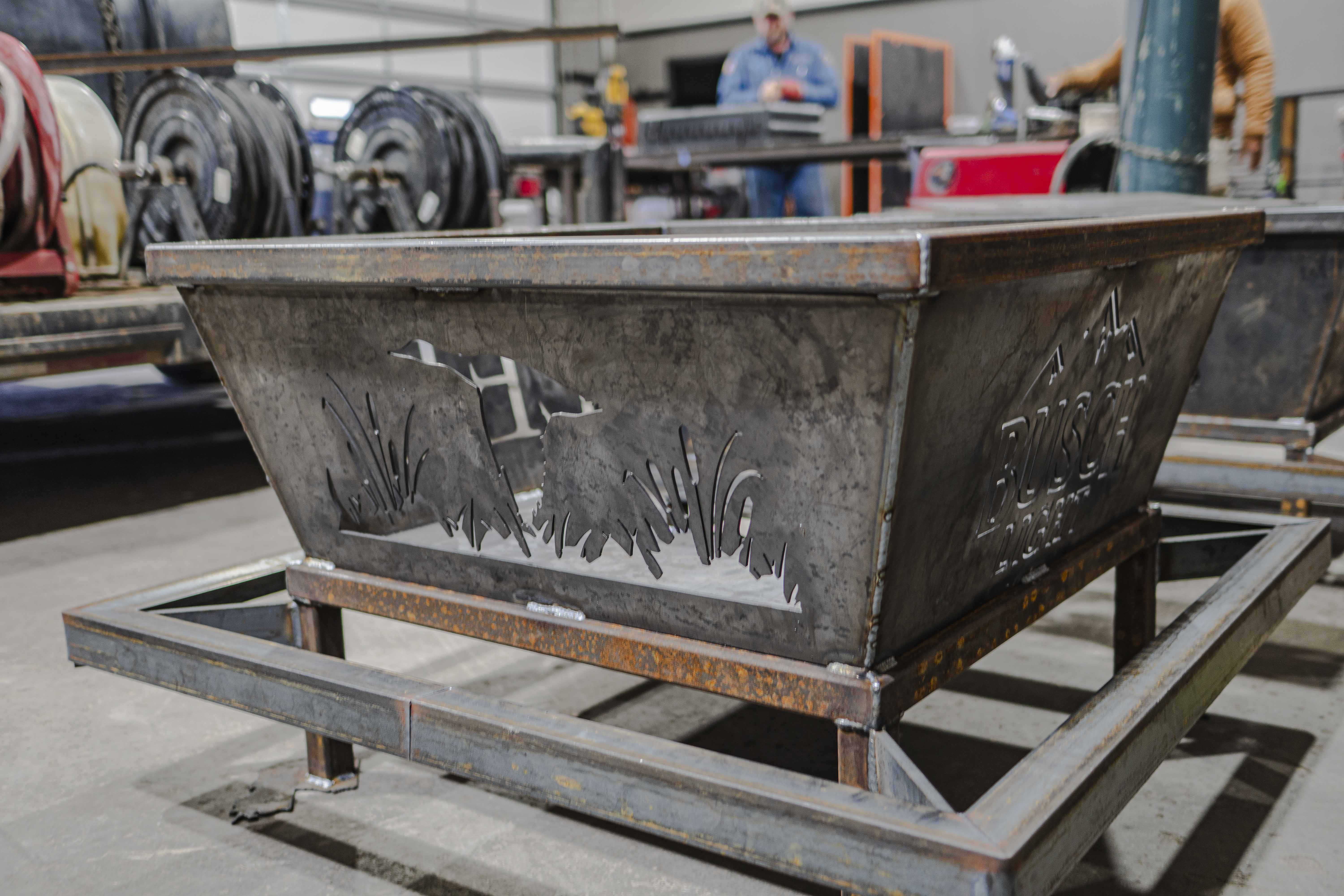 Rustic metal fire pit with cutout designs of a duck and grass, and the Busch Light logo on the side.
