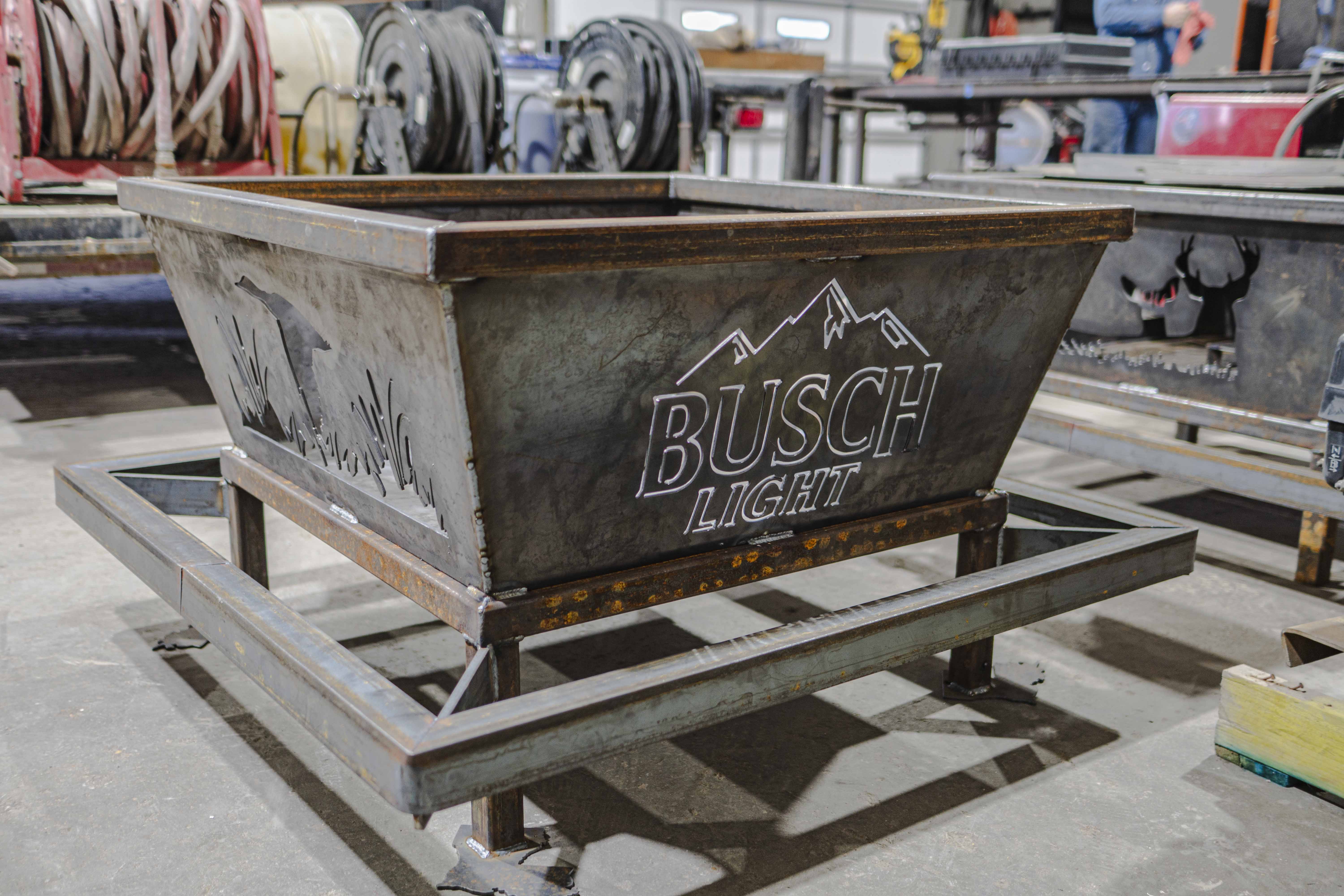 Metal fire pit with Busch Light logo and mountain design, placed on a square steel frame in a workshop.