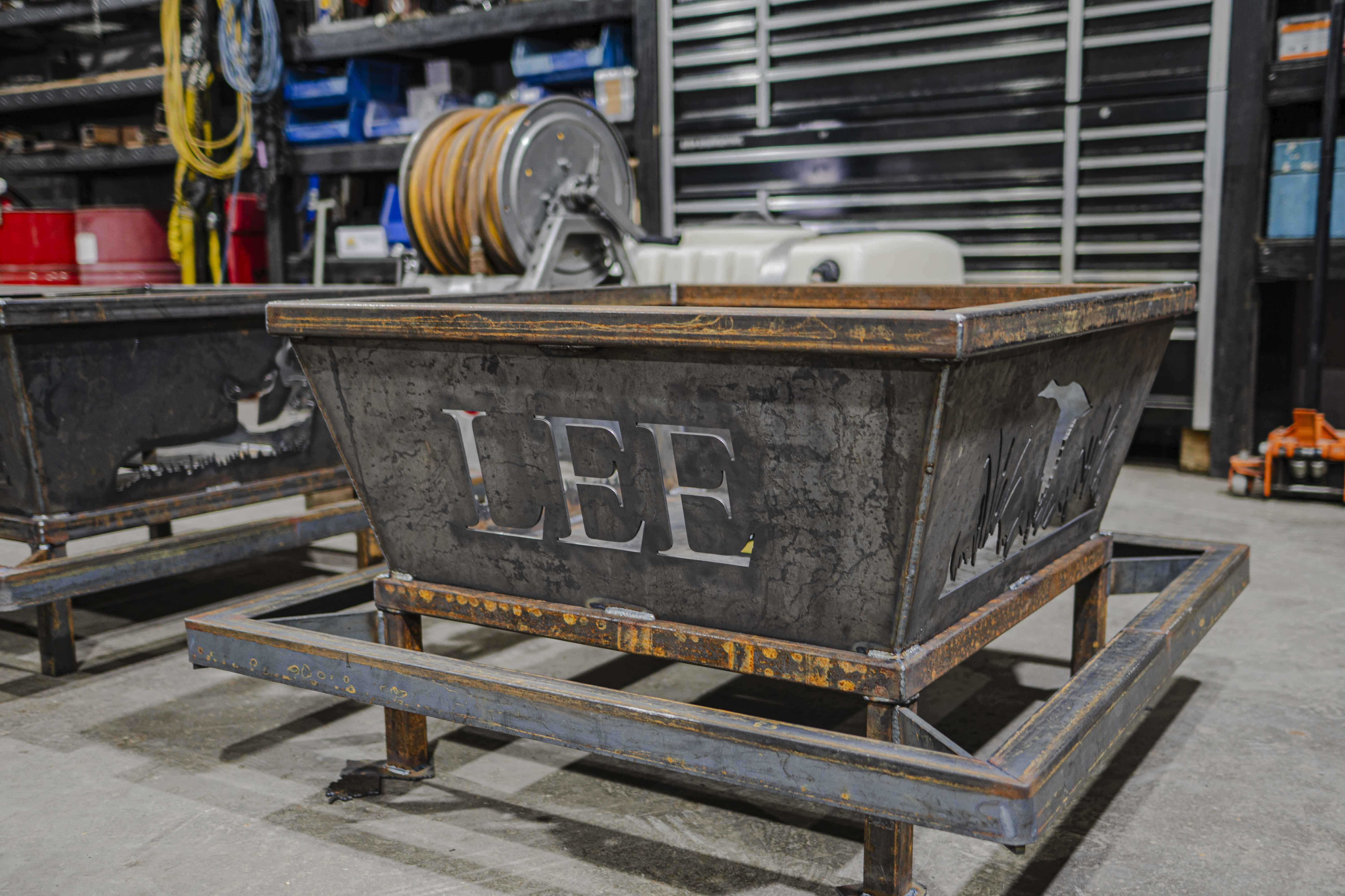 Rustic metal fire pit with cut-out letters 'LEE' and nature-themed designs, in a workshop setting.