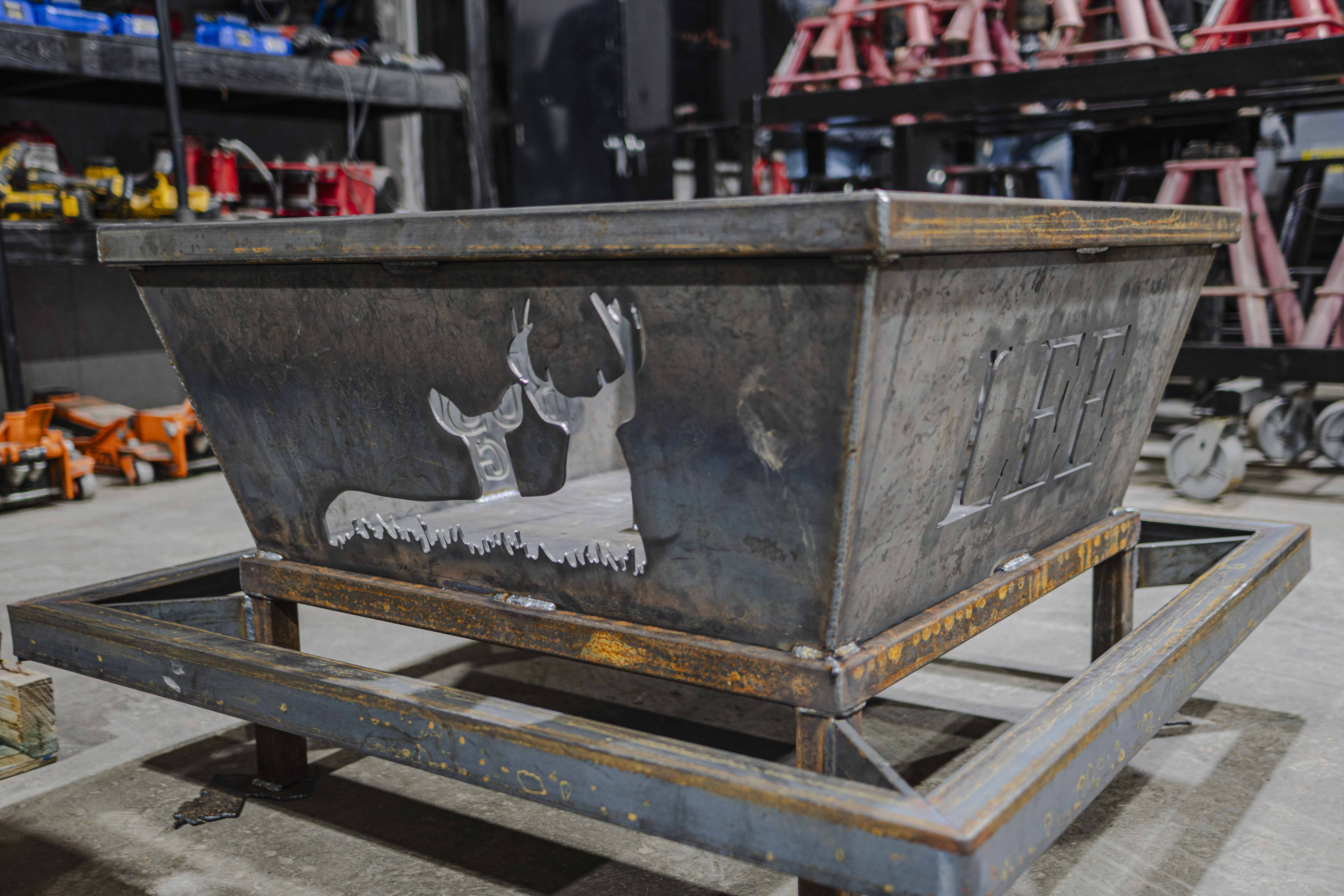 Rustic metal fire pit with cutout design of two deer heads and grass, placed in an industrial workshop setting.