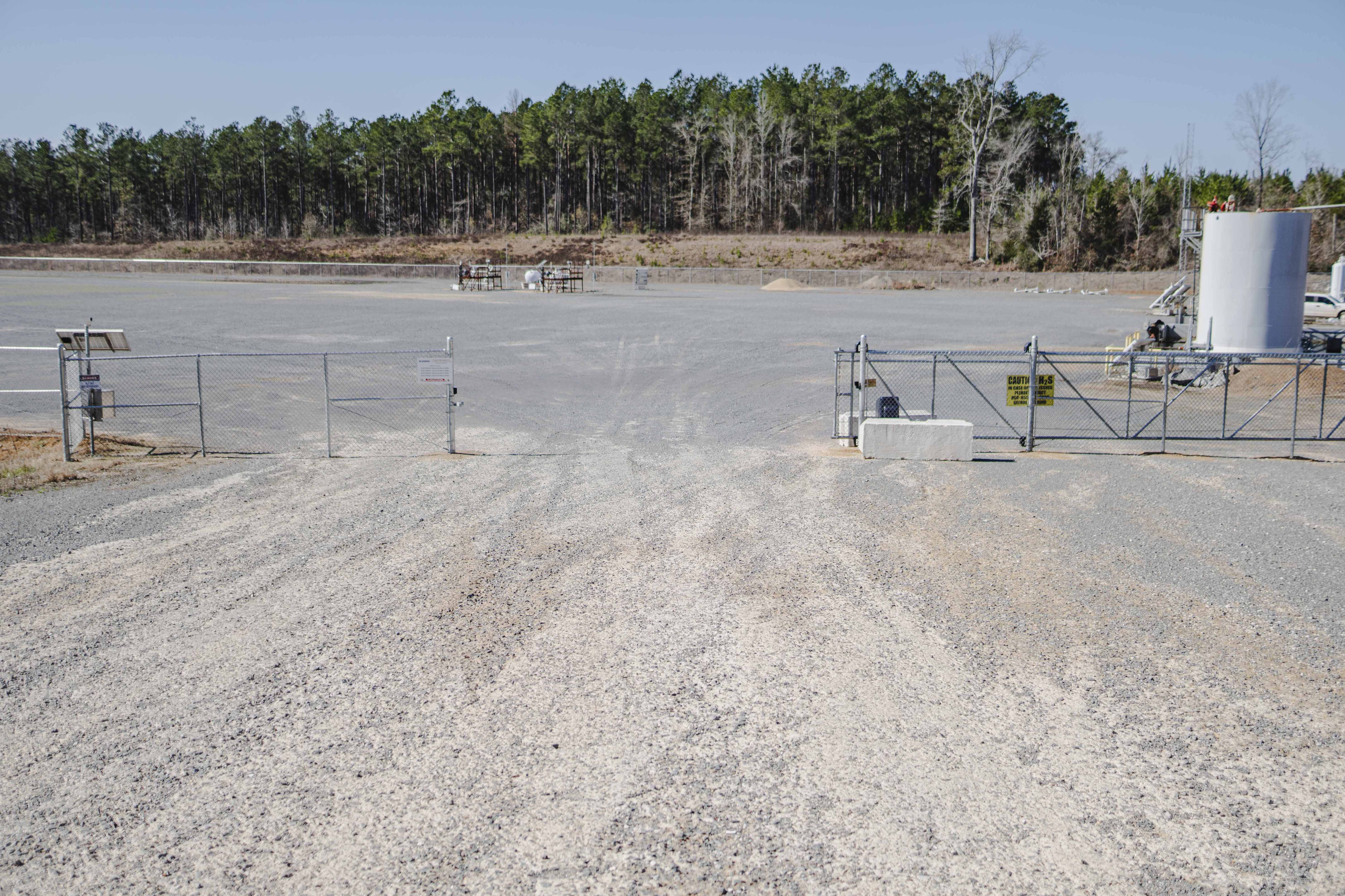 Gravel road leading to a closed metal gate securing an industrial site with storage tanks and equipment, backed by a forest.
