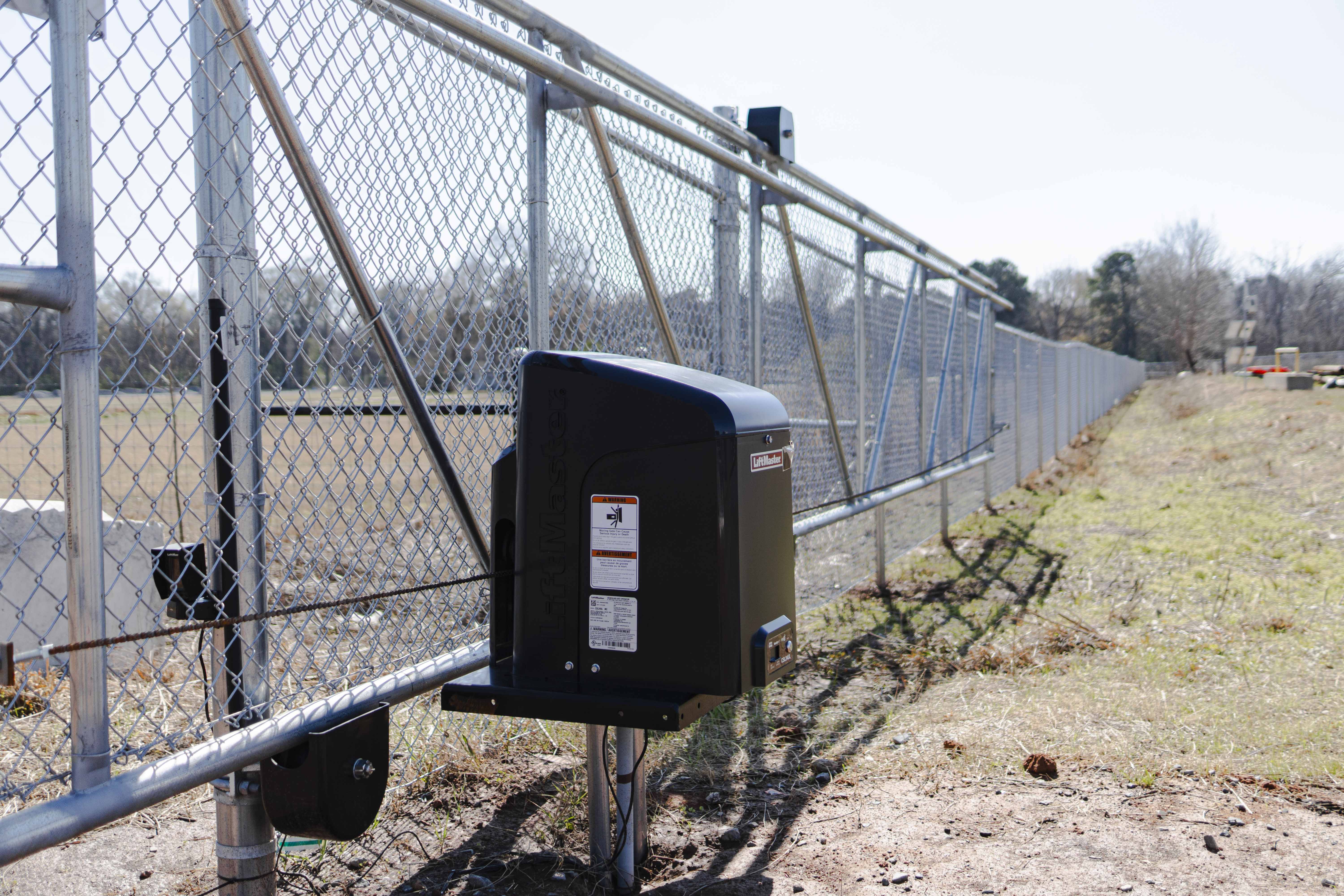 Automated metal sliding fence gate with a black LiftMaster motor and chain mechanism on a gravel and grass area.