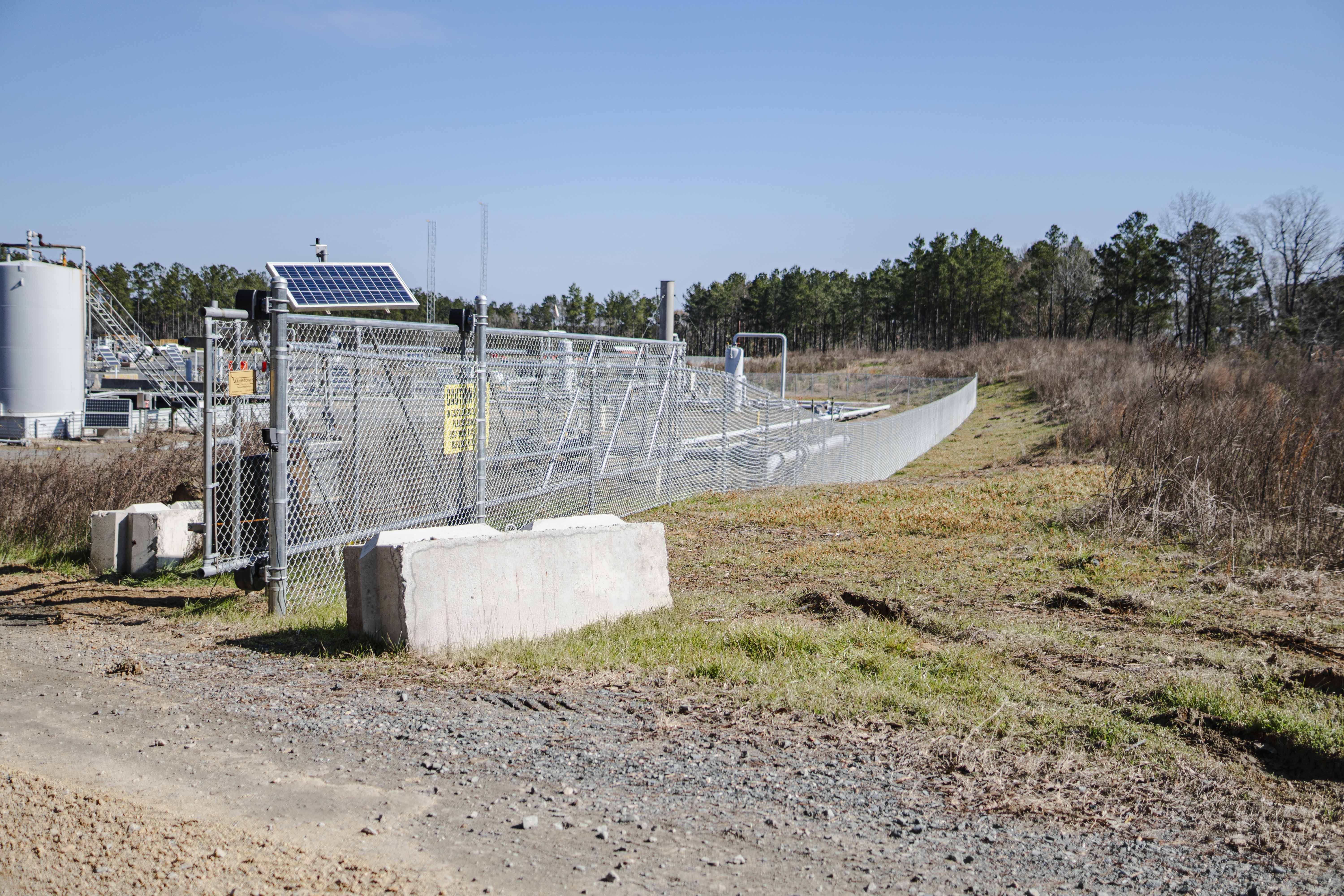 Chain-link fence with concrete barriers enclosing an industrial site with tanks and solar panels under a clear blue sky.
