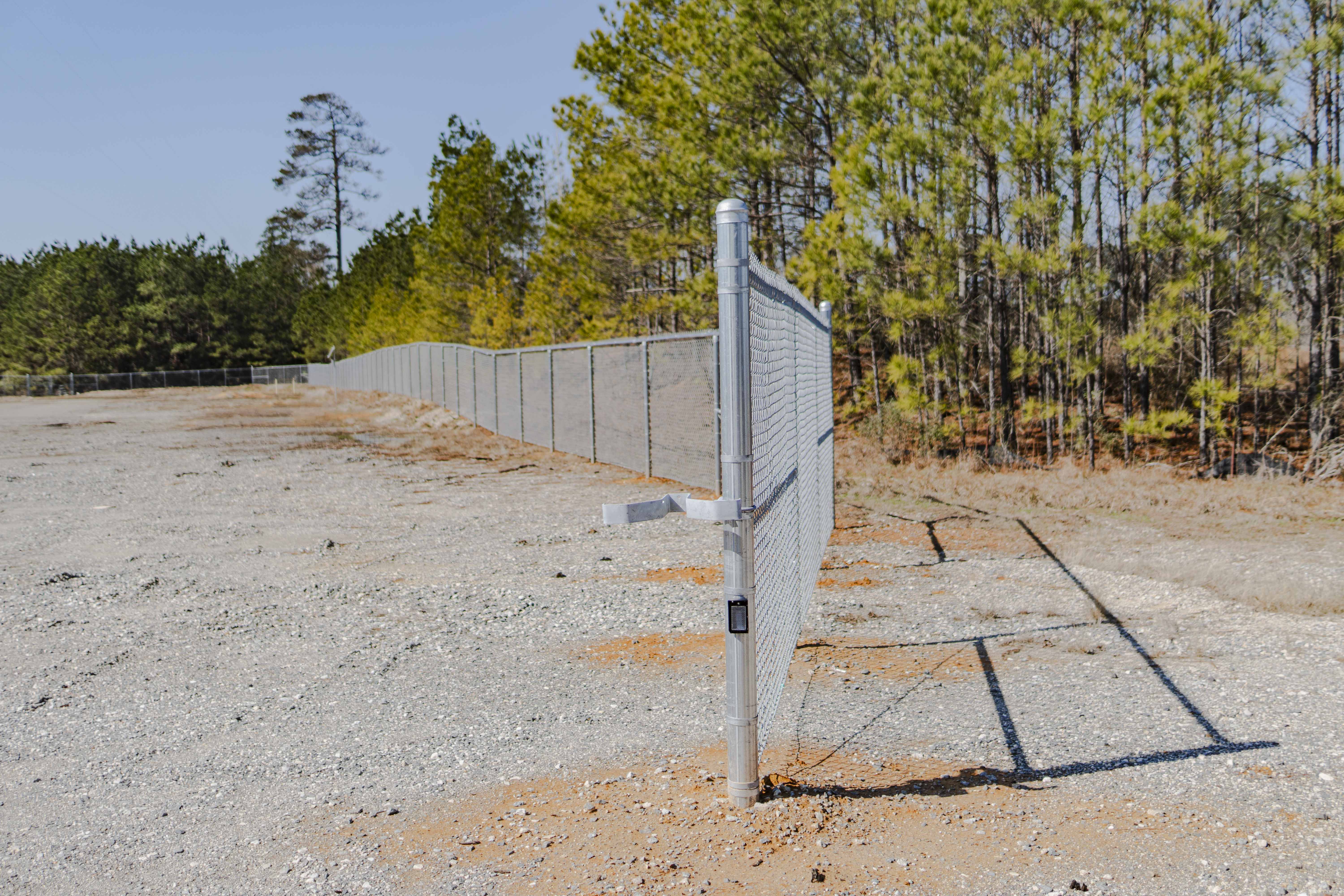 Long silver chain-link fence installed on a gravelly, dirt clearing with trees in the background under a clear sky.