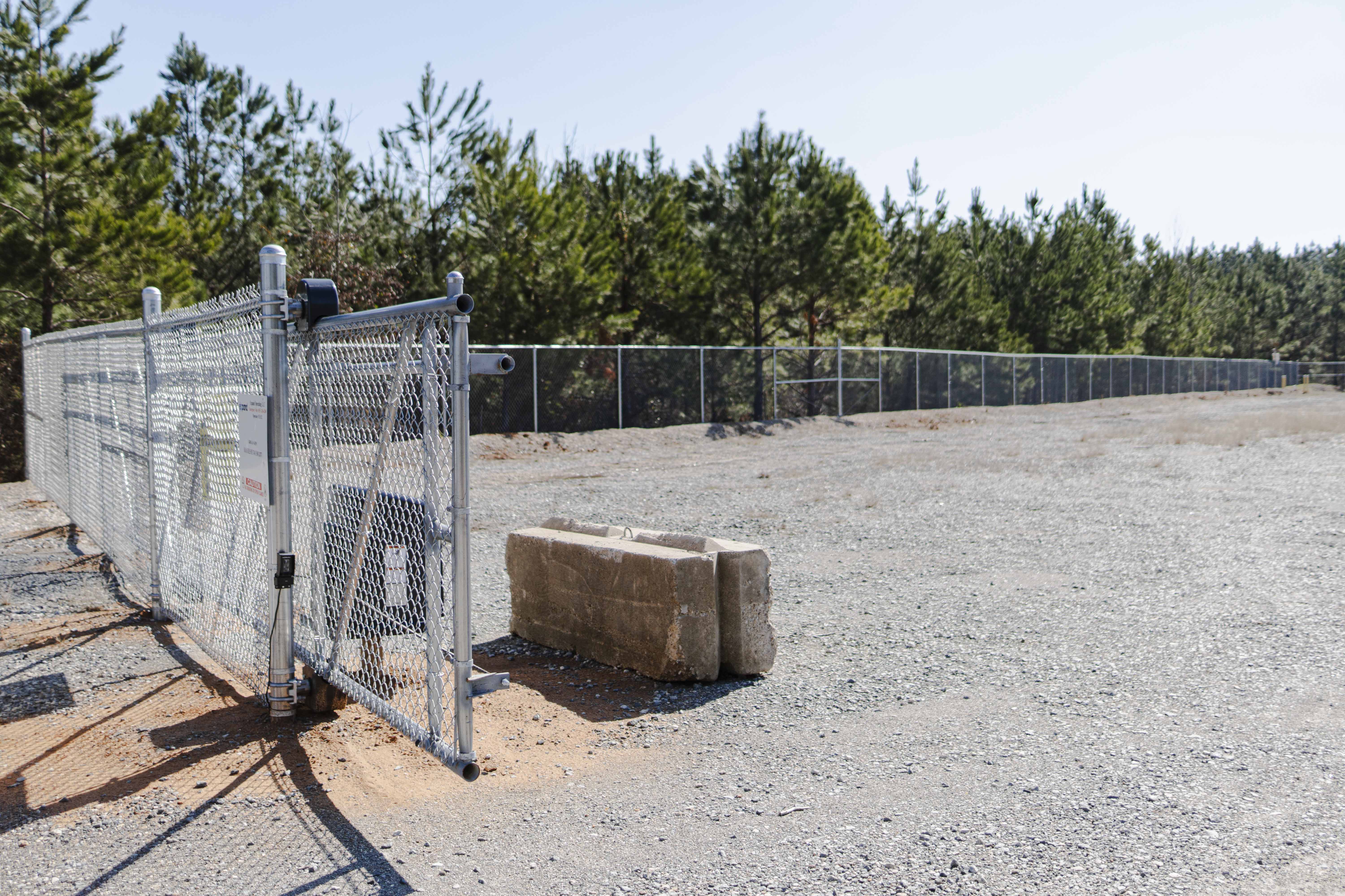 Opened chain-link security gate with concrete block barrier beside a gravel area and pine trees in the background.
