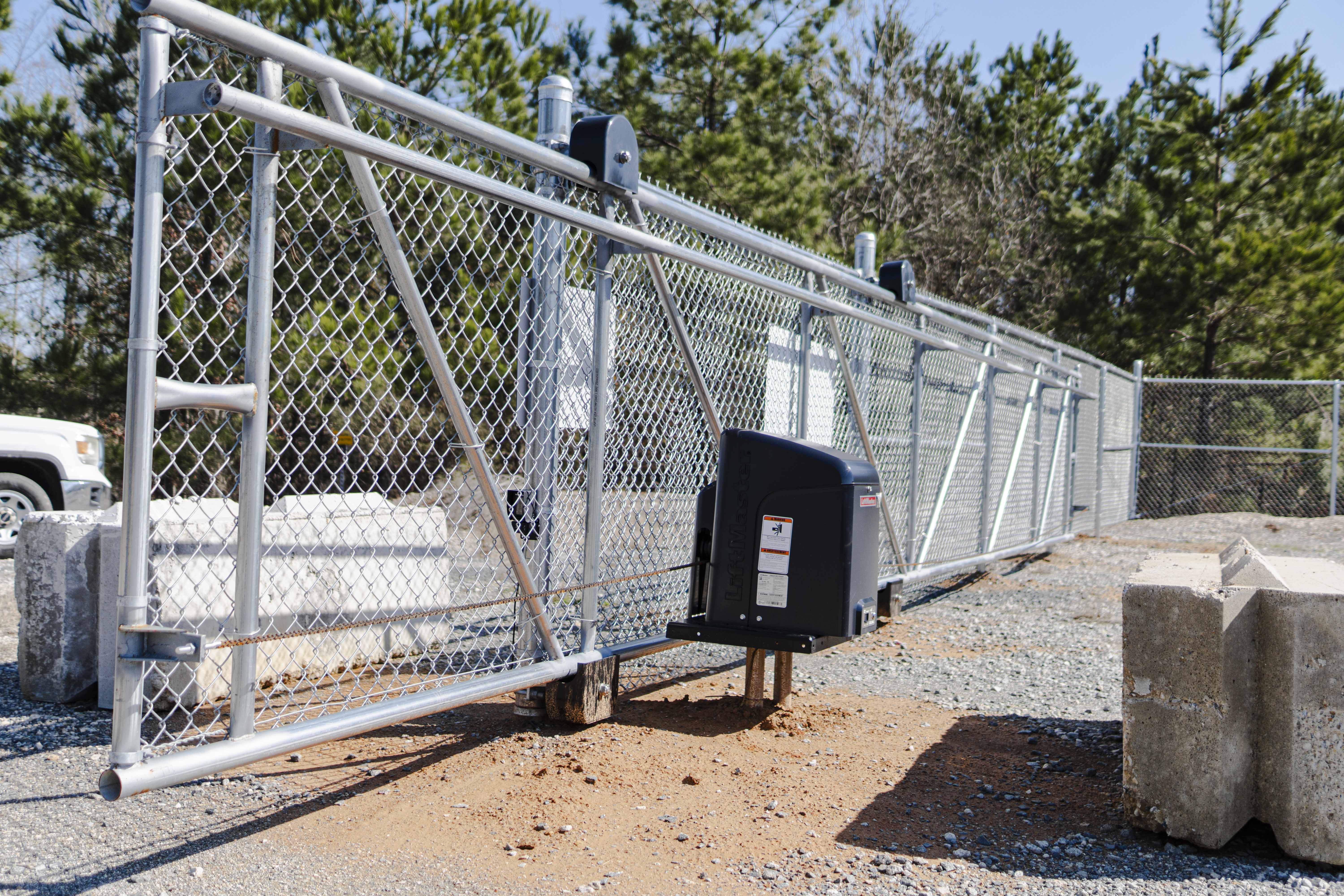 Metal chain-link sliding gate with an automatic gate opener attached, set against a background of trees and concrete barriers.