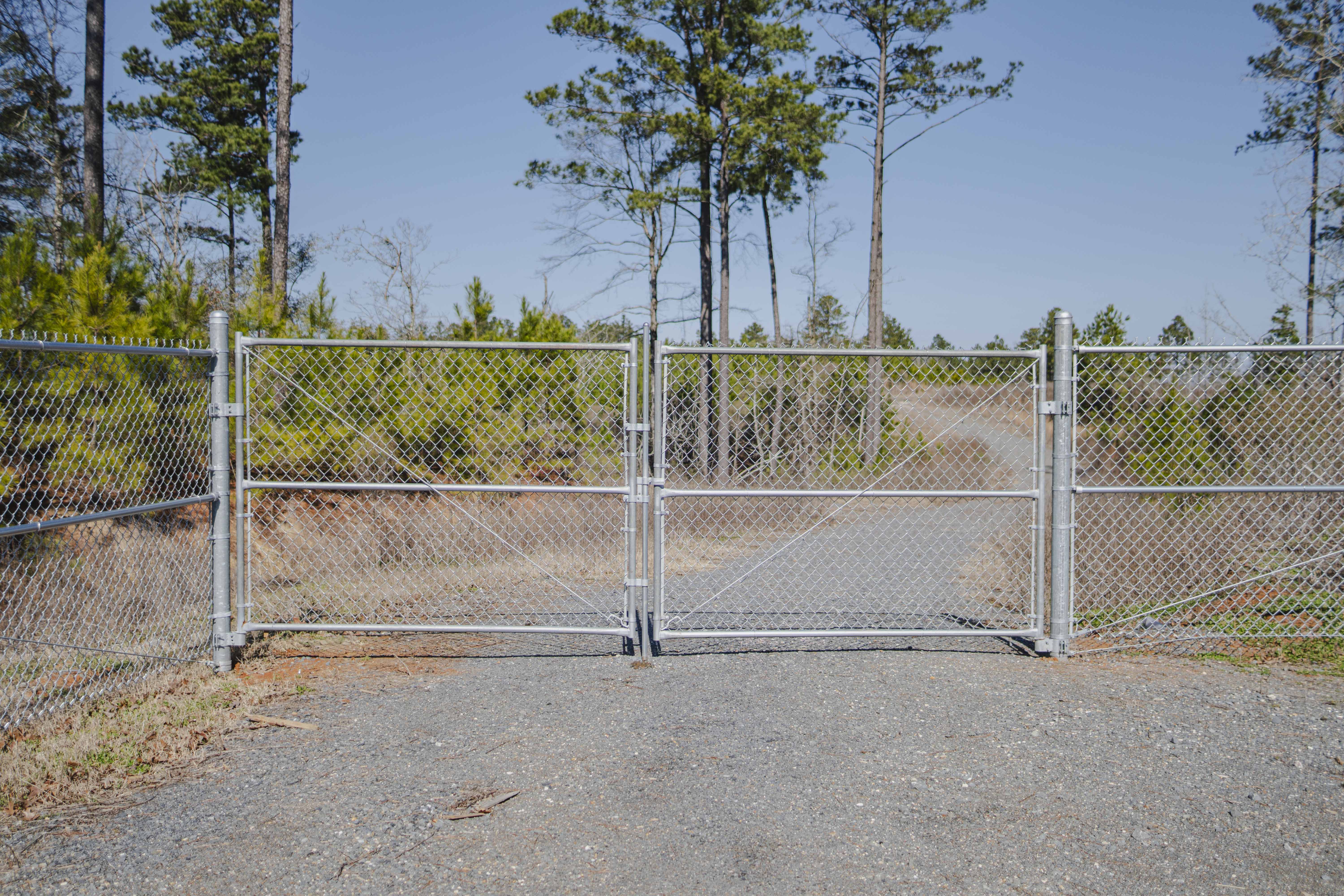 Metal chain-link gate blocking a gravel road leading into a wooded area under clear blue sky.
