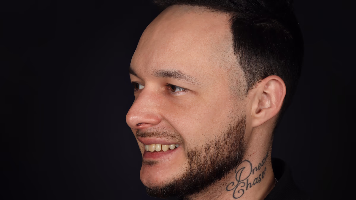 Портрет чоловіка з коротким темним волоссям, бородою та татуюванням на шиї на чорному фоні.