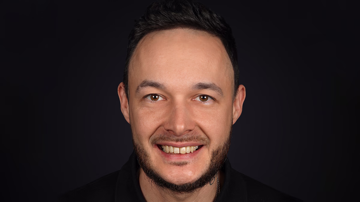 Чоловік із коротким чорним волоссям і короткою бородою усміхається на чорному фоні.