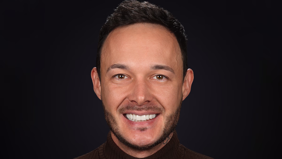 Чоловік зі світлою шкірою посміхається на чорному тлі, має коротке темне волосся і бороду.