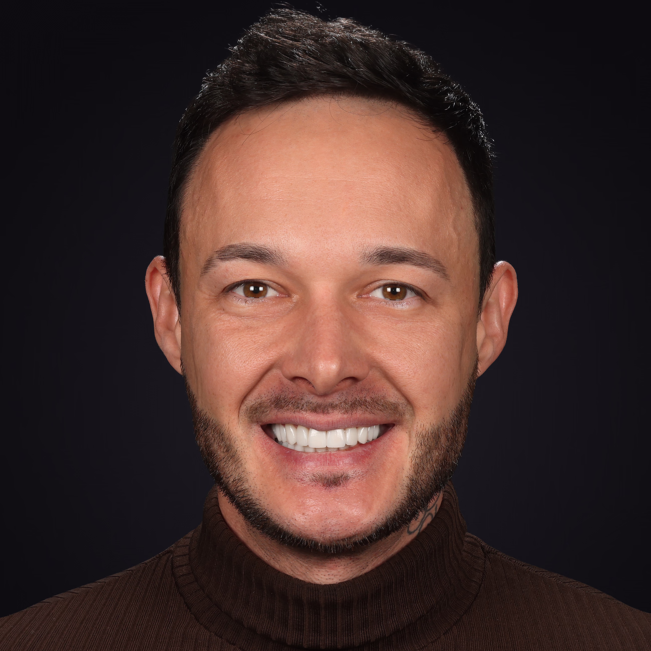 Чоловік із коротким темним волоссям і бородою посміхається, одягнений у коричневий гольф на темному фоні.