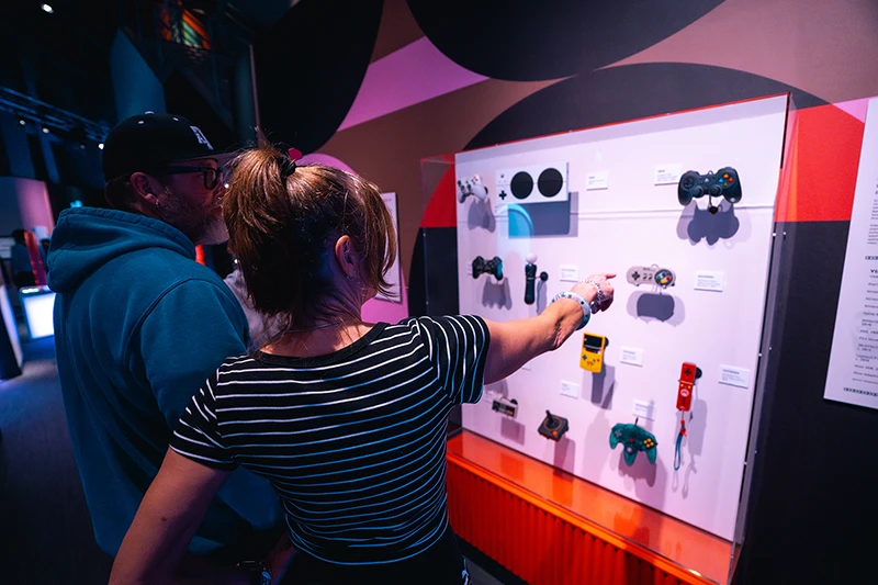 A young white woman points to video game artifacts in the Massive exhibition. An older white man stands beside her, looking at the artifacts