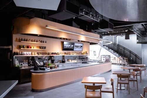 Modern cafe counter with light wood finishes, coffee machines, and snacks, with tables and chairs in an industrial-style interior.