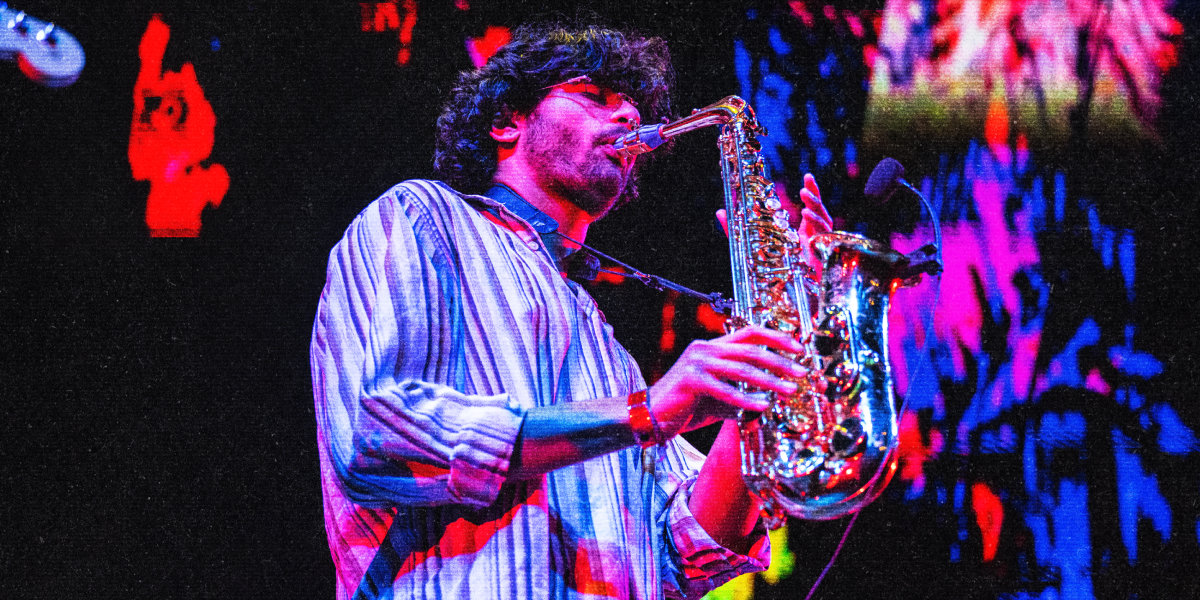 Man with curly hair and glasses playing a saxophone on stage with colorful abstract lights in the background.